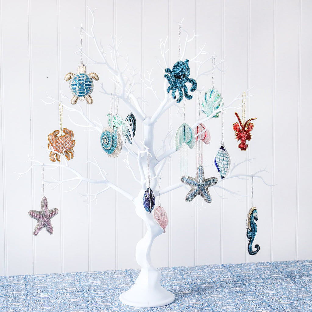 The image shows a white decorative tree with various sea-themed beaded ornaments hanging from its branches. The ornaments include a turtle, an octopus, a crab, a seashell, a starfish, a seahorse, and other marine creatures. Each ornament is intricately crafted with small beads in different colors like blue, green, red, and gold. The tree is set on a blue patterned tablecloth, with a plain white wall as the backdrop. The scene has a festive, coastal, or nautical holiday theme.