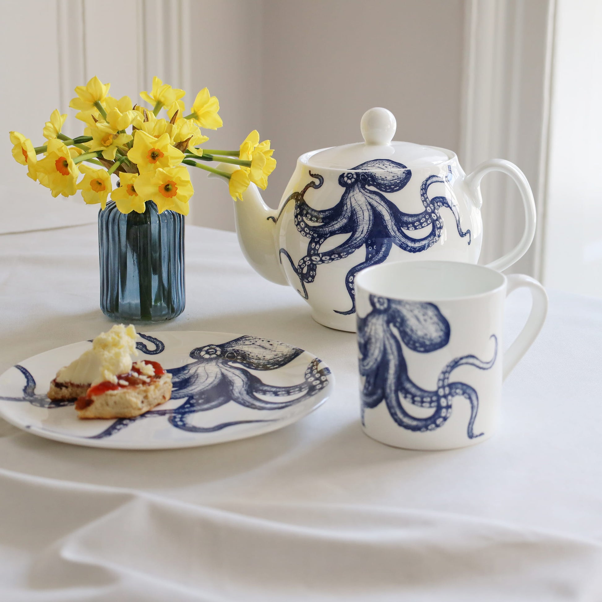 Tea set with blue octopus design on a white tablecloth, accompanied by a plate of food and a vase of yellow flowers.