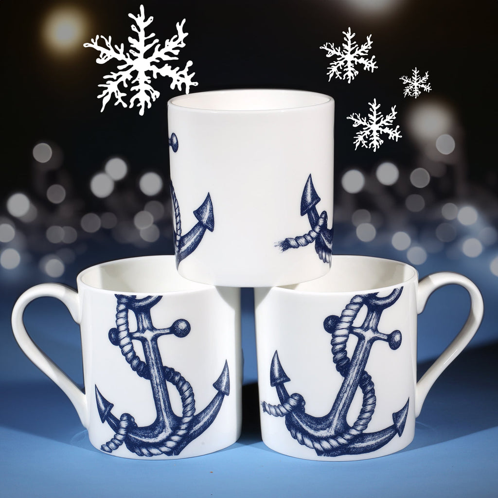 Three white mugs featuring detailed navy blue anchor illustrations arranged in a pyramid shape, with softly glowing lights and delicate white snowflakes in the background, creating a festive nautical winter theme.