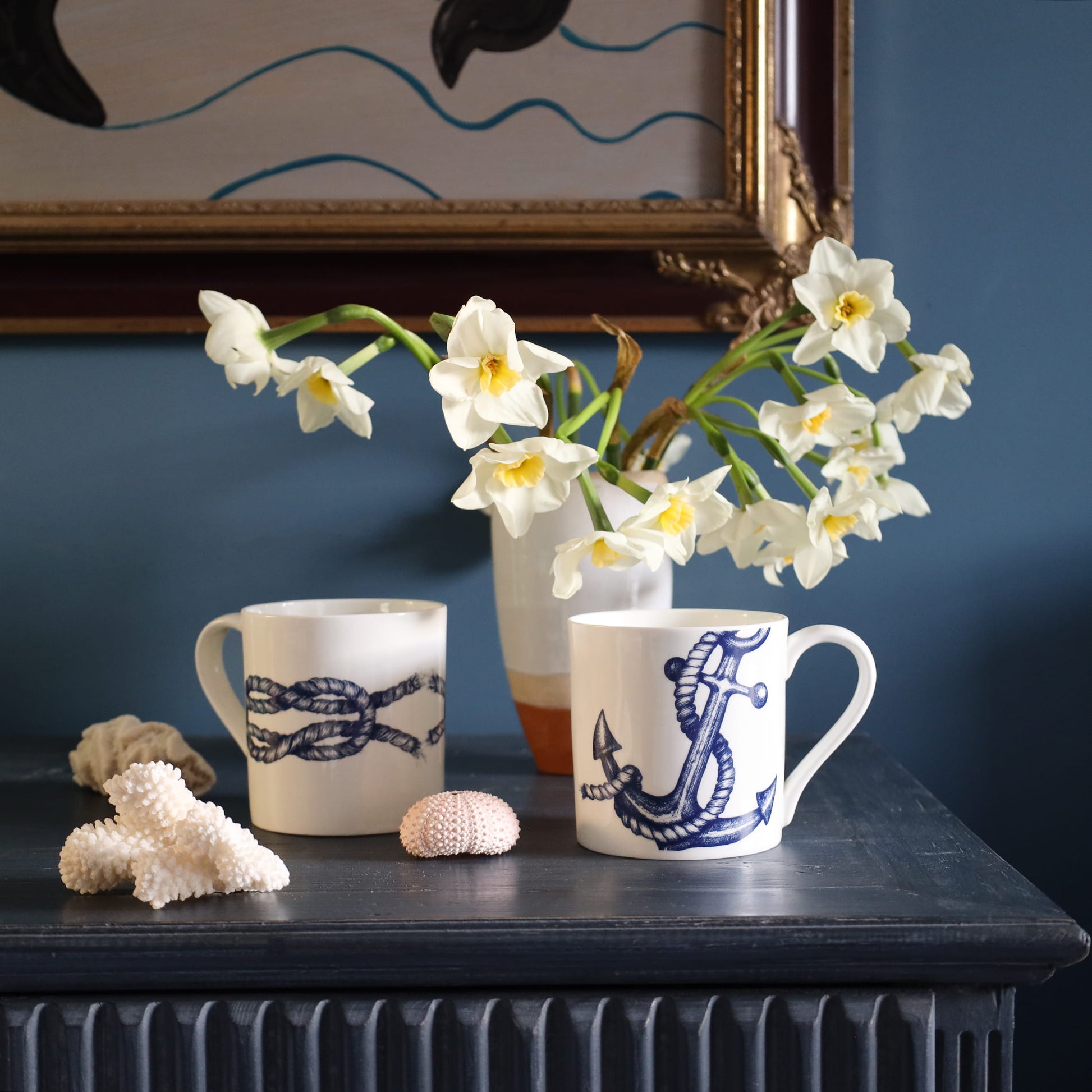 Two mugs with nautical designs on a surface with flowers and shells.