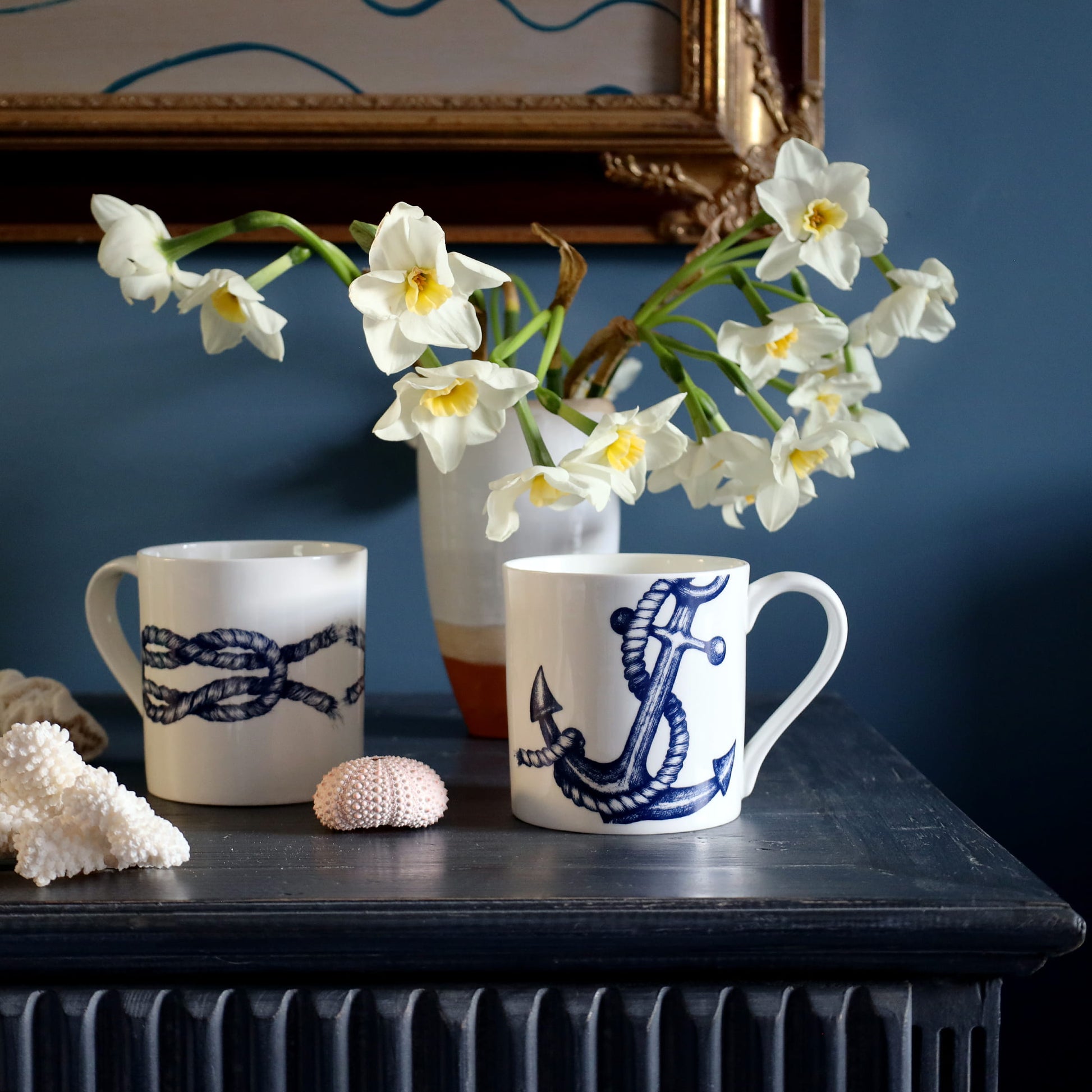 Two ceramic mugs with anchor & knot design on a dark surface with flowers in the background.