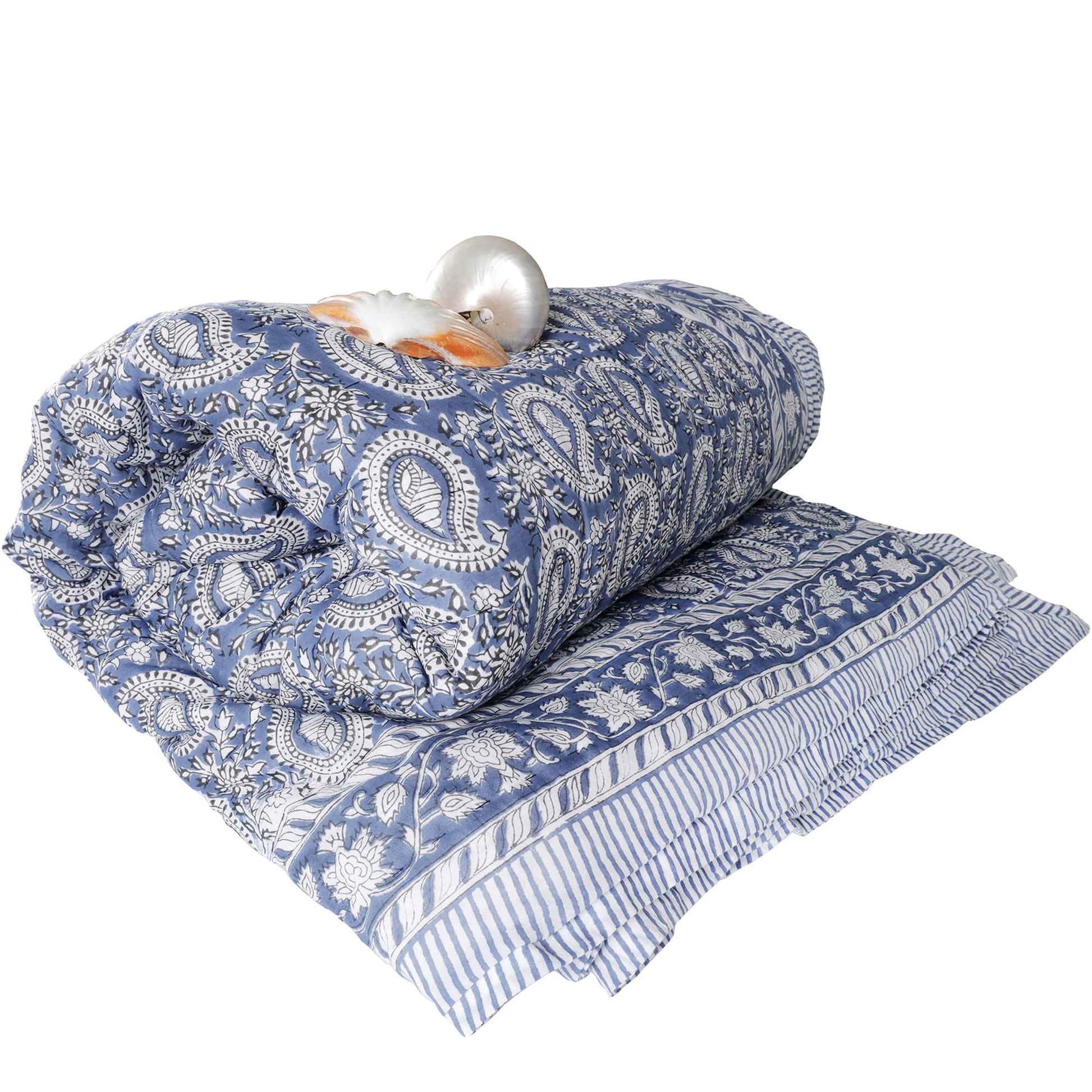 Rolled blue quilt with intricate paisley and floral patterns, accented by striped edges. A decorative seashell and a nautilus shell are placed on top for added aesthetic