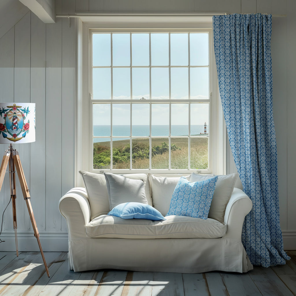 A bright coastal living room with a large window overlooking the sea and a distant lighthouse. The space features a white sofa with blue and grey cushions, including one with a coral-inspired pattern that matches the curtain beside the window. A tripod lamp with a nautical-themed shade stands to the left, completing the serene seaside interior.