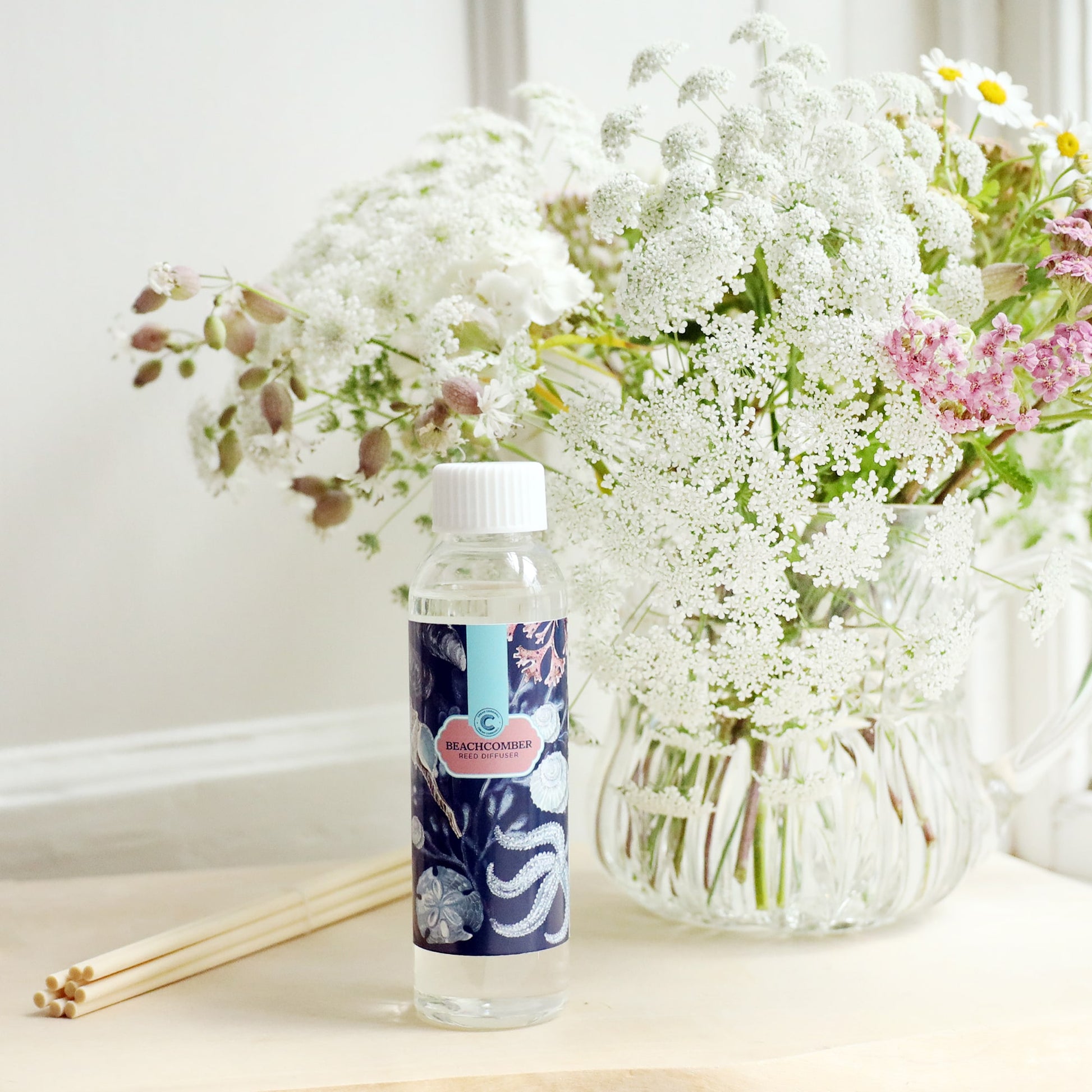 Bottle of Beachcomber room diffuser oil with octopus design on the label, sitting on a wooden block with the diffuser reeds laying jut to the side, and a vase of soft coloured flowers sitting behind.