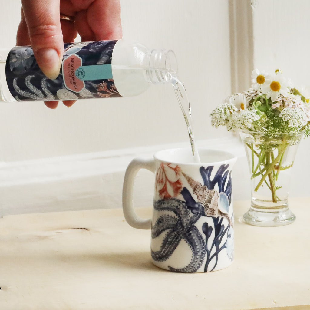 Mini Jug with beachcomber design on with reeds and small posy of flowers behind with a bottle of Beachcomber room diffuser oil being poured into the jug.