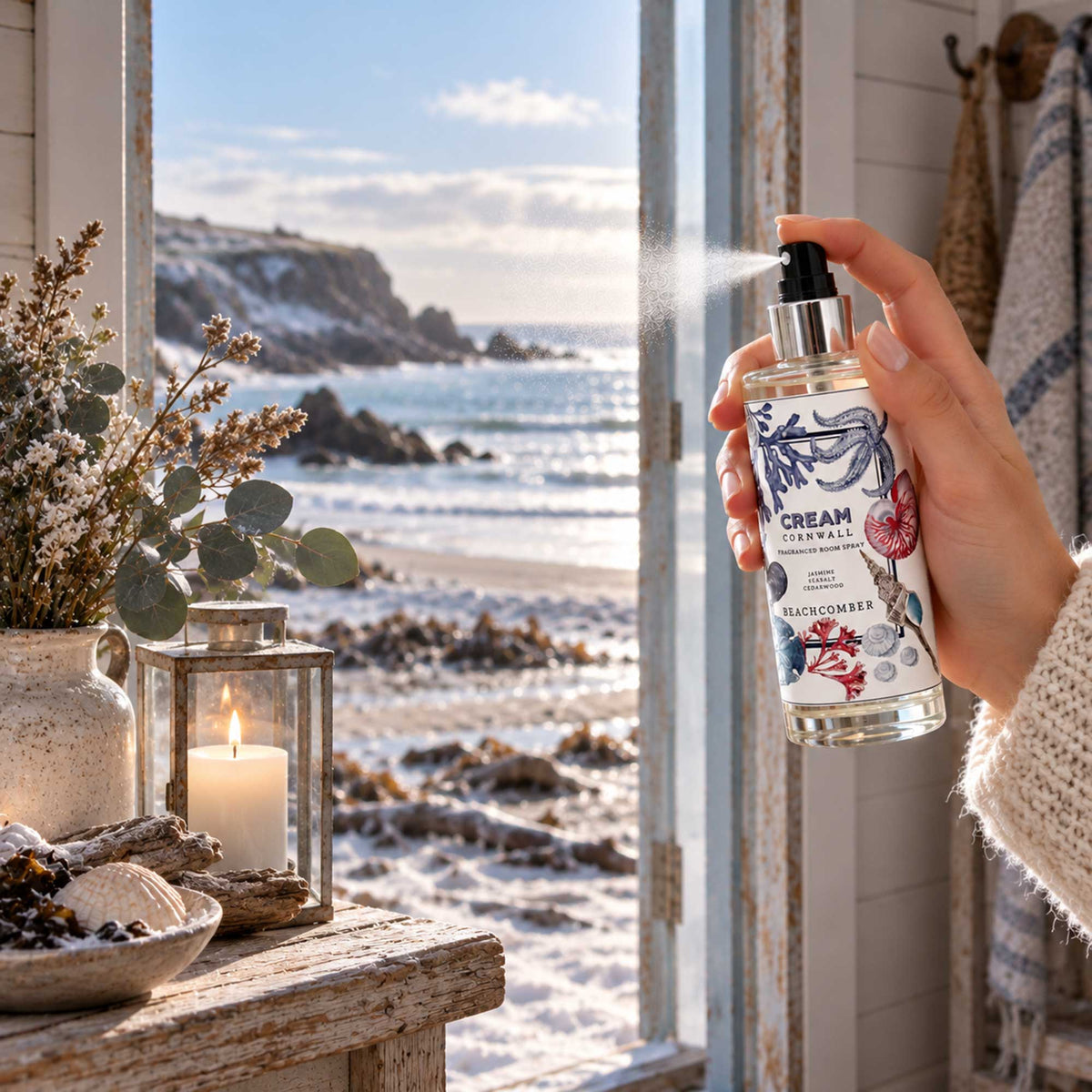 Hand spraying Cream Cornwall Beachcomber fragranced room spray in a cosy coastal cottage, with a lit candle, seashells, driftwood and a sunlit Cornish beach visible through the open doorway.