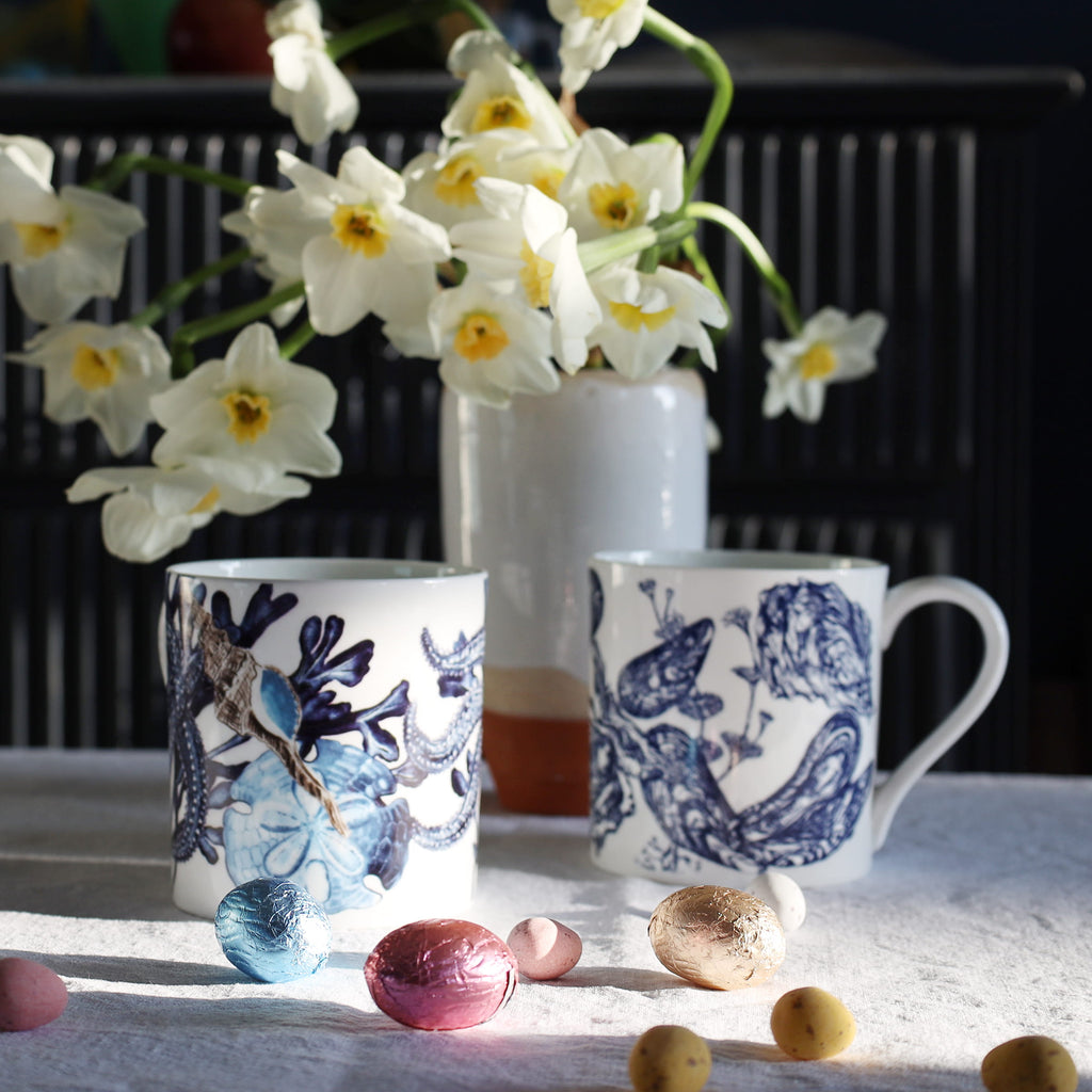 Two blue and white mugs with beachcomber designs on a table with flowers and Easter eggs.