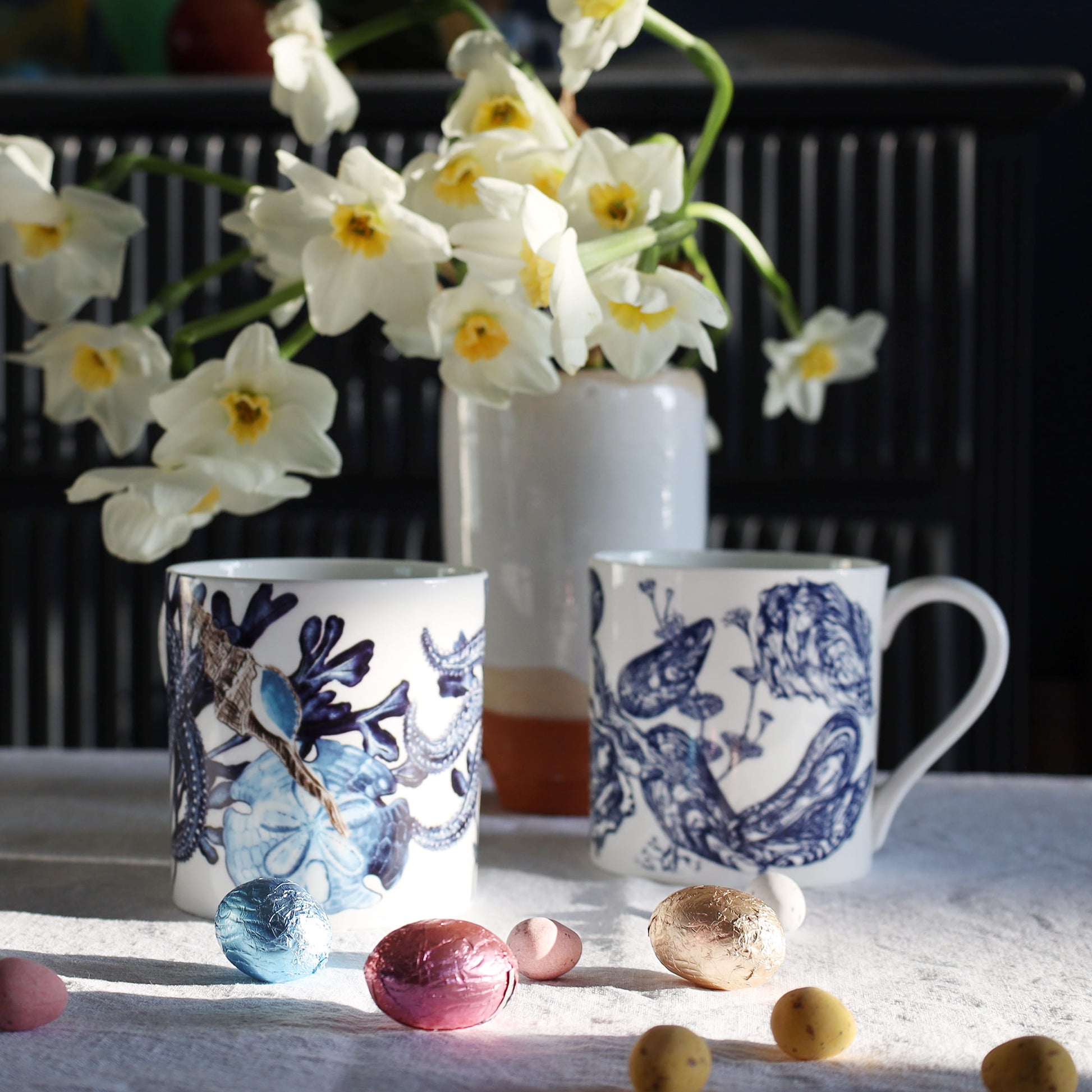 Two blue and white mugs with beachcomber designs on a table with flowers and Easter eggs.