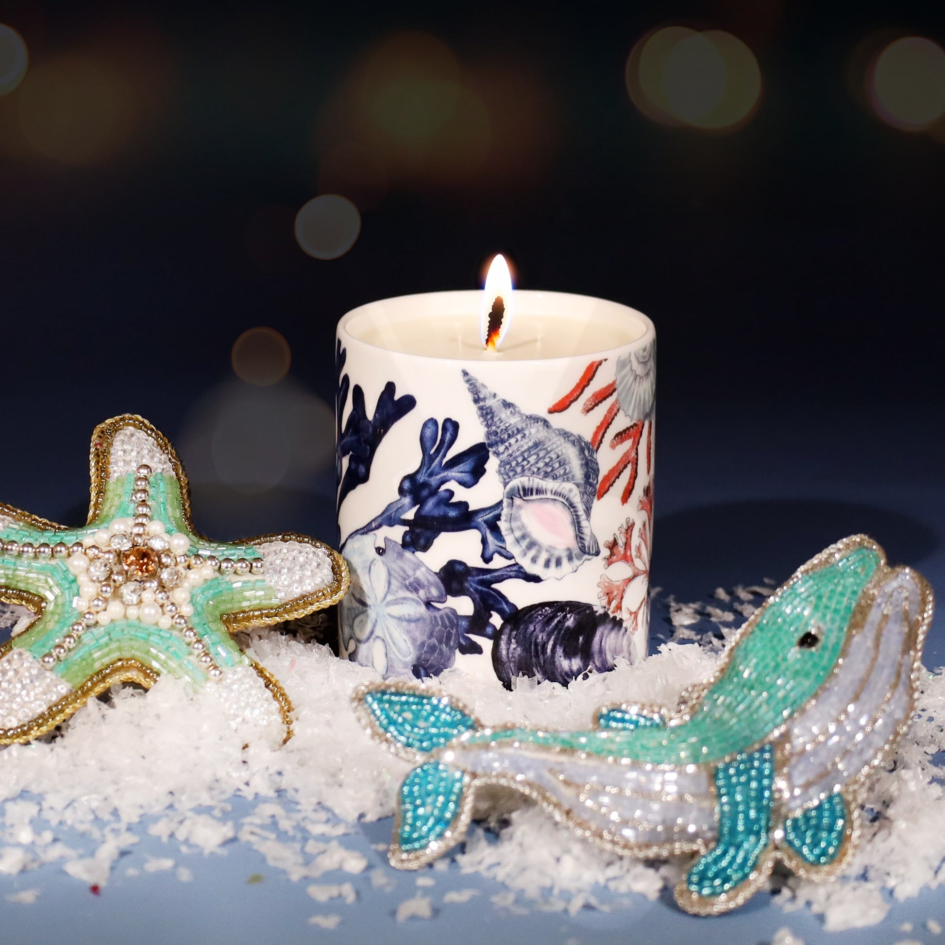Lit candle in a white holder decorated with blue and coral seashell and seaweed illustrations, surrounded by faux snow and beaded marine ornaments shaped like a starfish and whale, set against a dark festive background with warm golden lights.