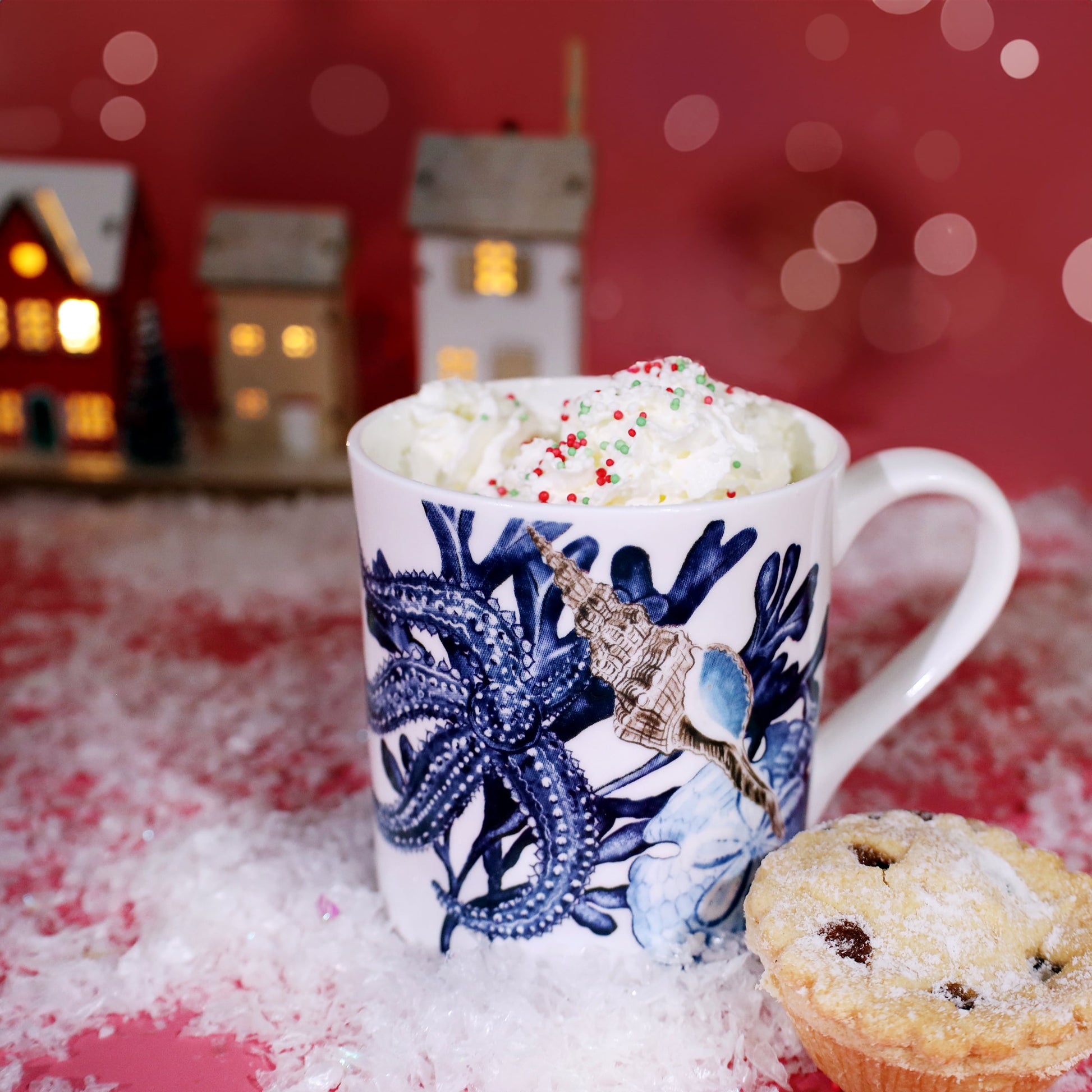 Festive scene featuring a white mug decorated with blue starfish and seashell illustrations, filled with whipped cream and colorful sprinkles, set on faux snow with a mince pie beside it and miniature lit Christmas houses in the background against a warm pink backdrop.