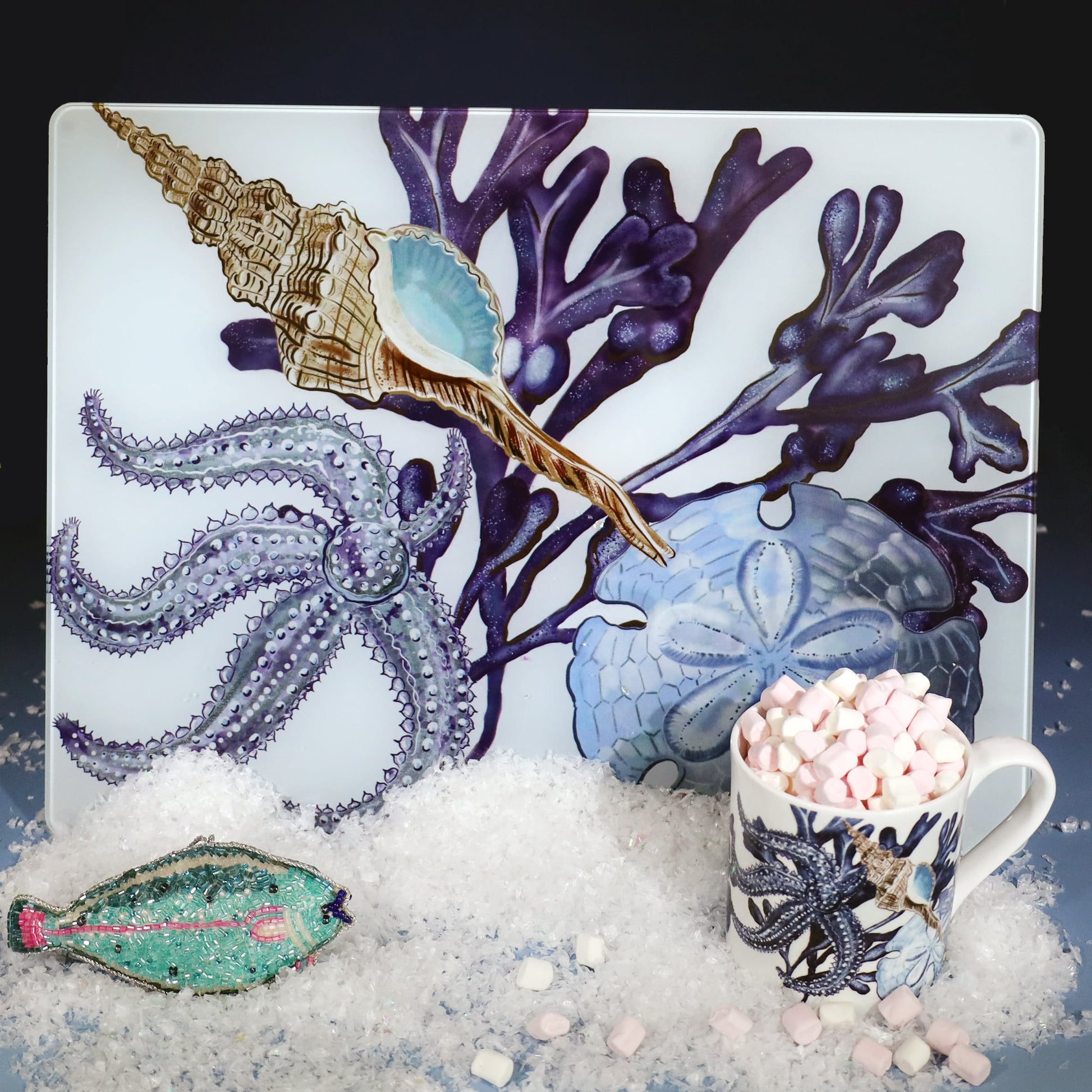 Beachcomber worktop saver featuring detailed illustrations of coral, shells, and a starfish in shades of blue and purple, displayed on faux snow beside a matching mug filled with marshmallows and a beaded fish ornament.
