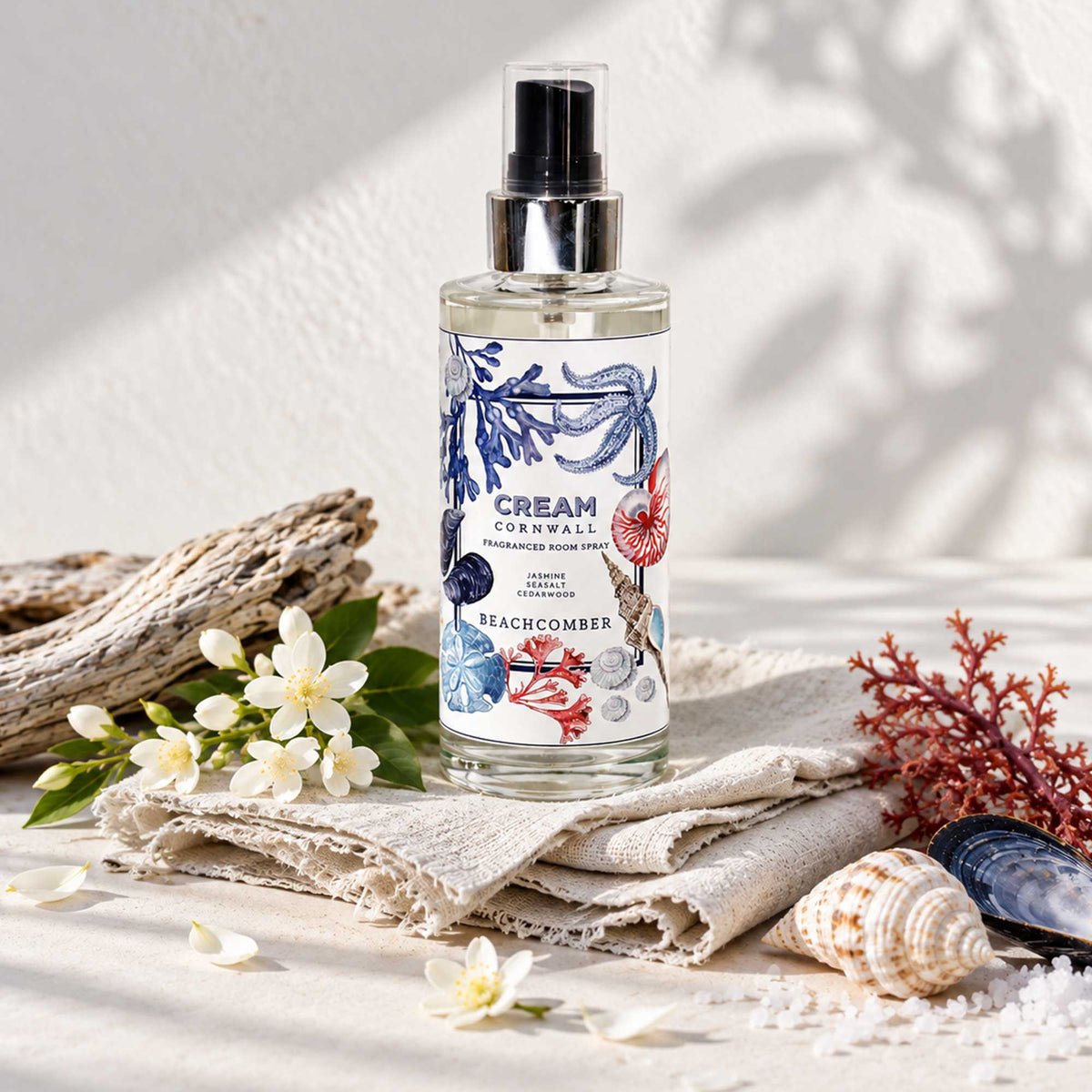 Cream Cornwall Beachcomber fragranced room spray with coastal-inspired botanical and seashell illustrations, displayed on natural linen alongside white jasmine flowers, driftwood, seashells, sea salt and red coral against a softly sunlit neutral background.