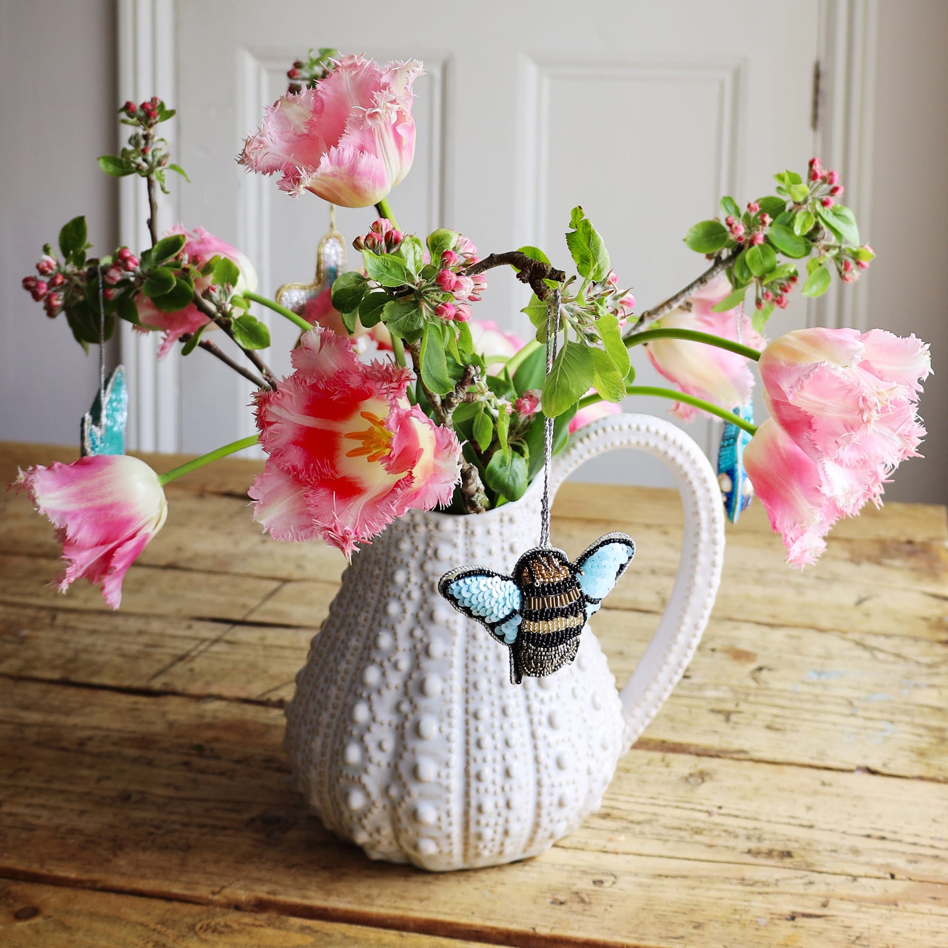 Urchin jug with a beaded bee decoration hanging from pink flowers on a wooden surface.