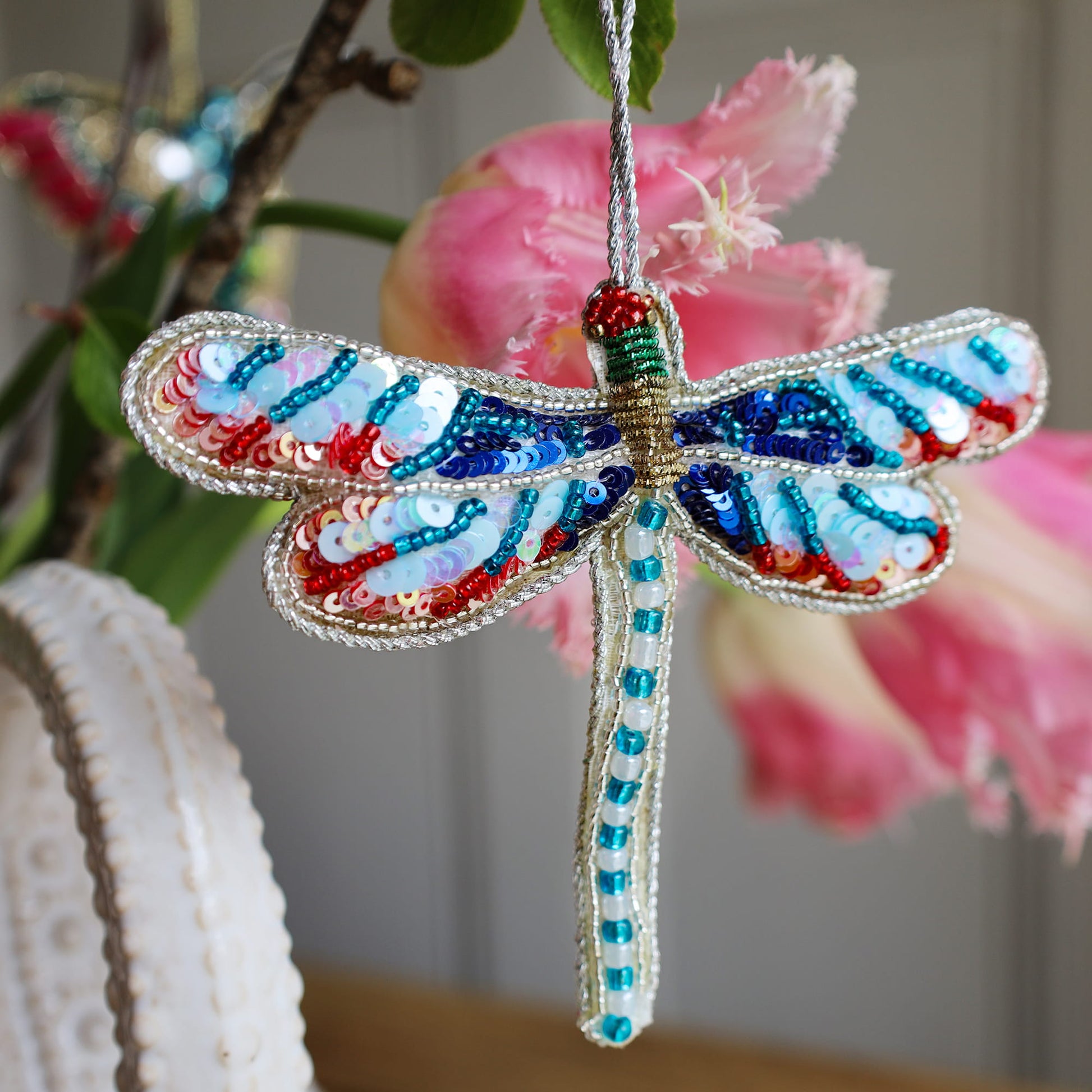Decorative dragonfly ornament with beads and sequins, hanging against a blurred floral background.