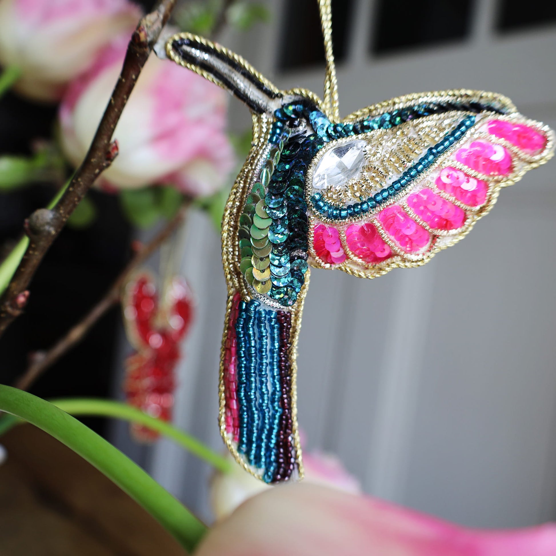 Colorful beaded hummingbird ornament held against a blurred floral background