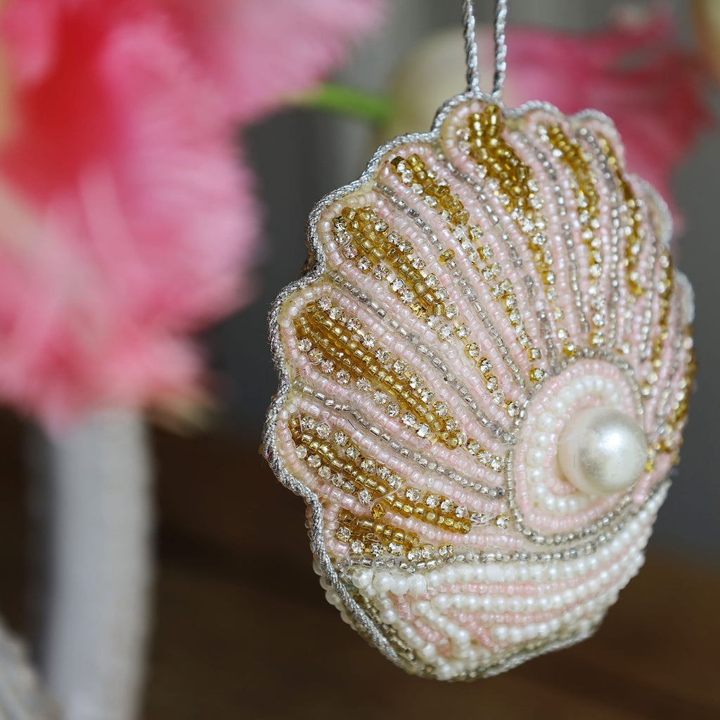 Decorative shell ornament with beads and a pearl, blurred pink flowers in the background