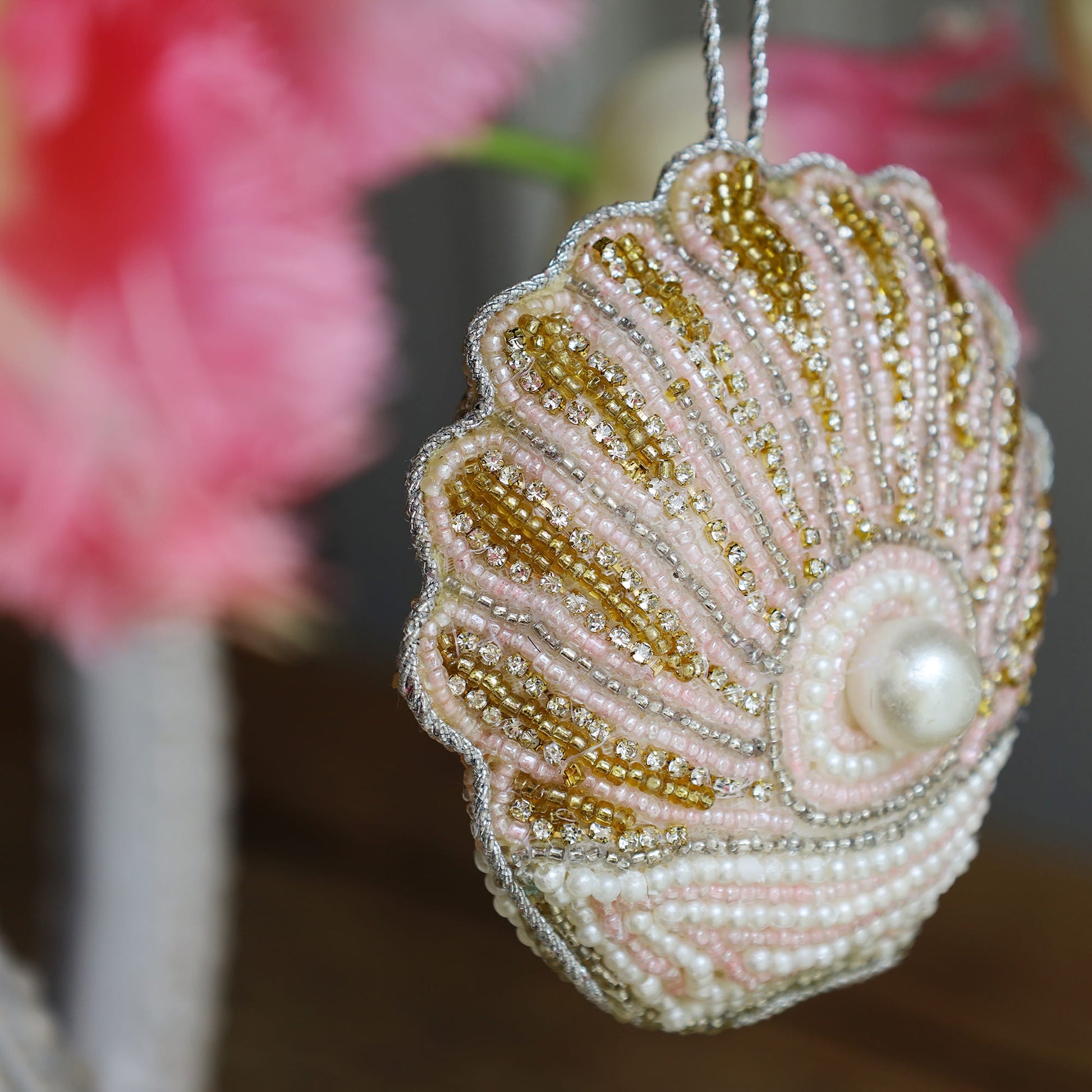 Decorative shell ornament with beads and a pearl, blurred pink flowers in the background