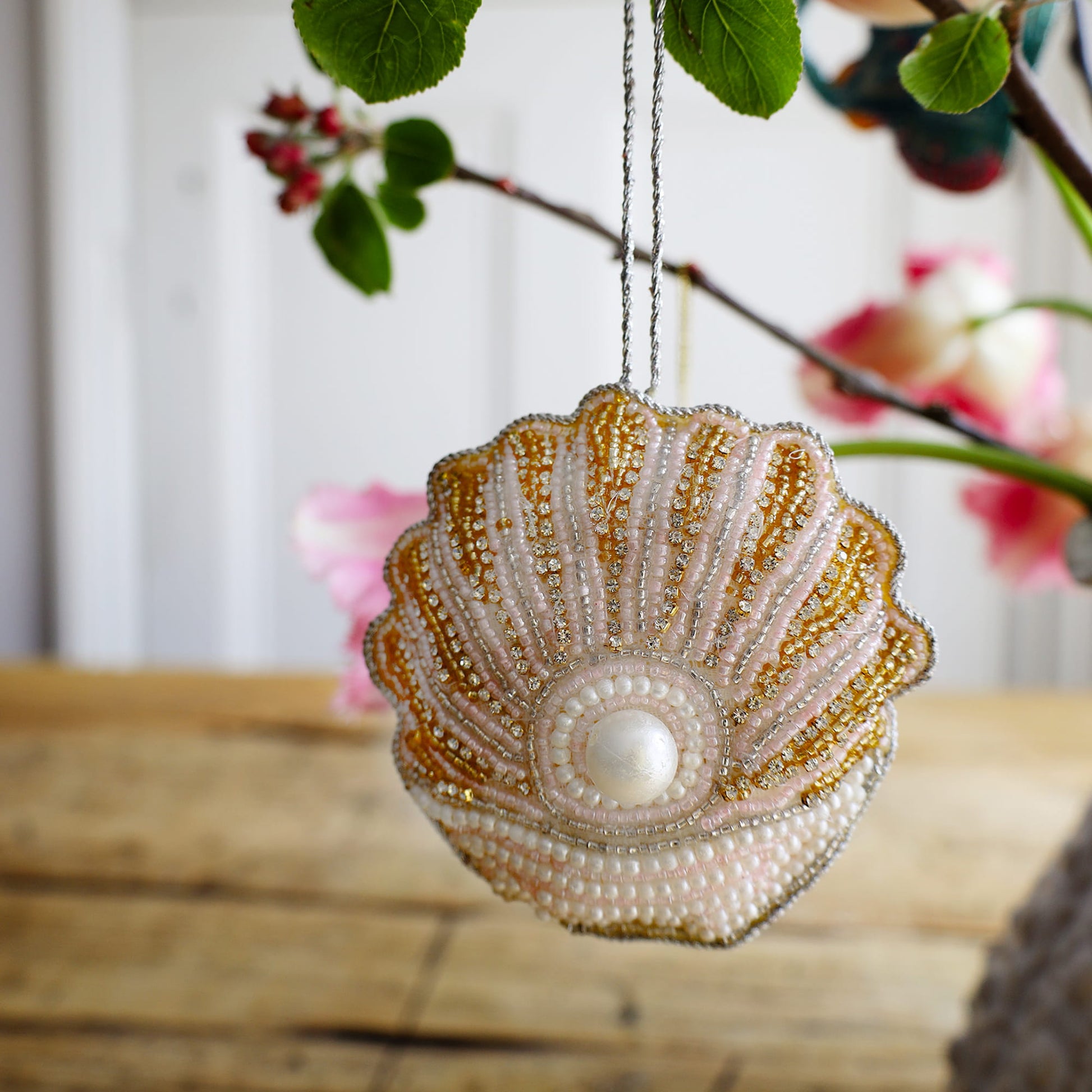 Decorative shell ornament with beads and a pearl on a wooden surface with blurred floral background