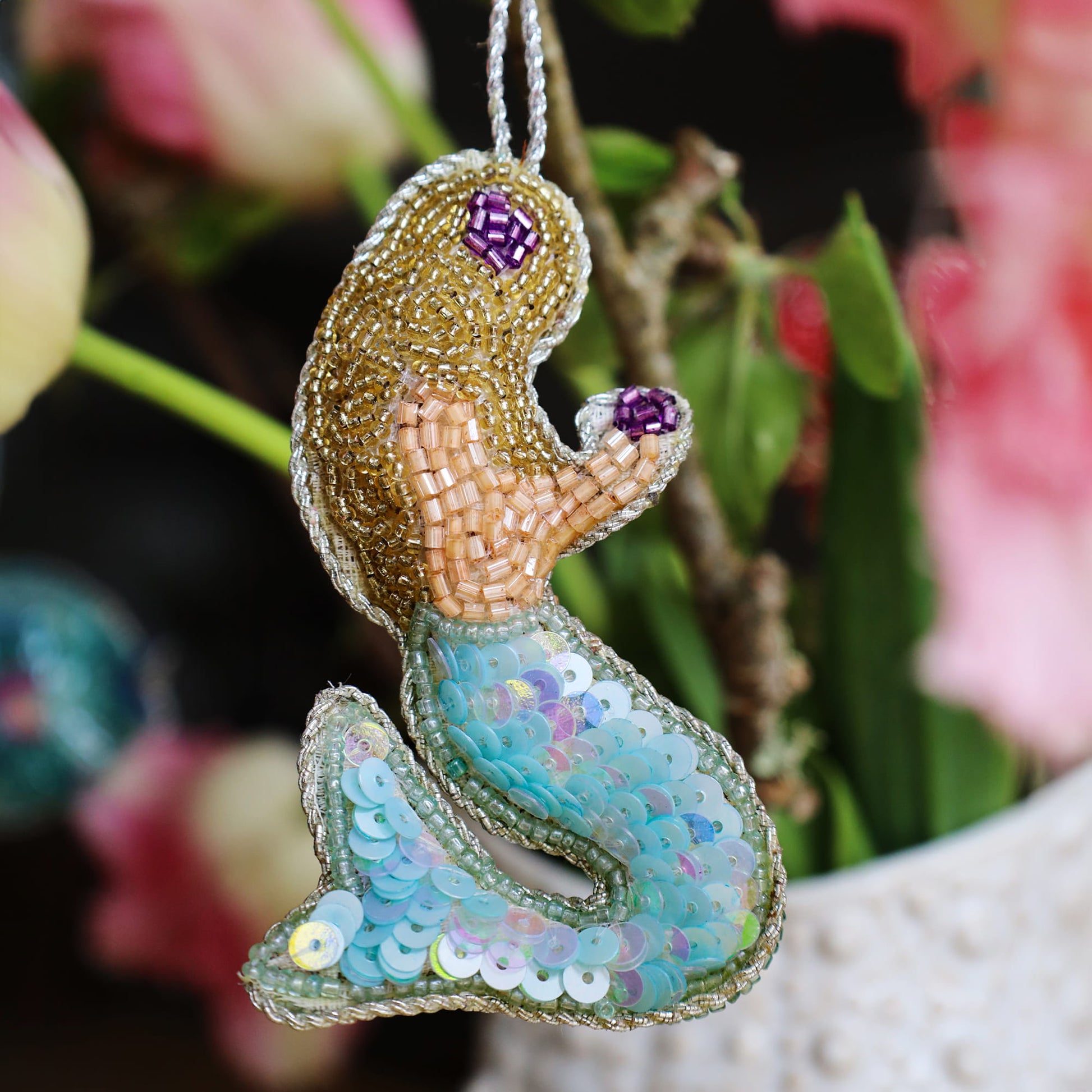 Decorative mermaid ornament with sequin tail and beaded top against a floral background
