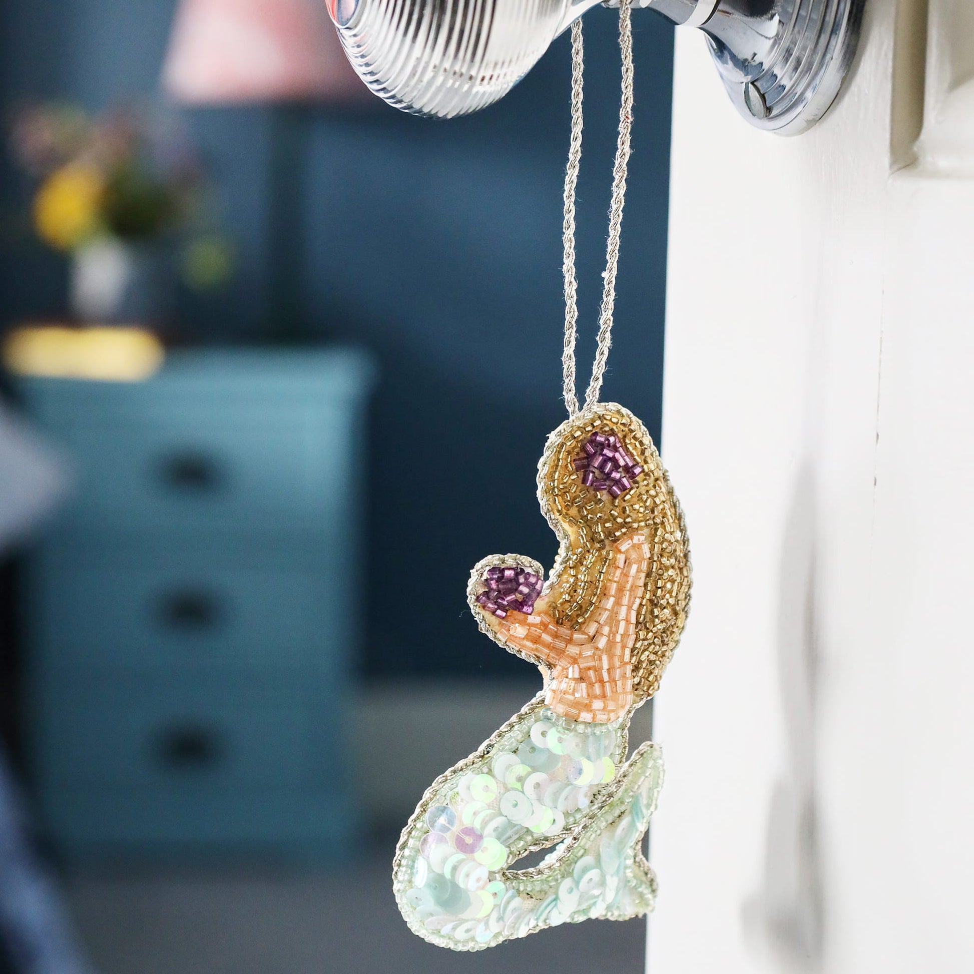 Decorative mermaid tail ornament hanging on a hook with a blurred room background.
