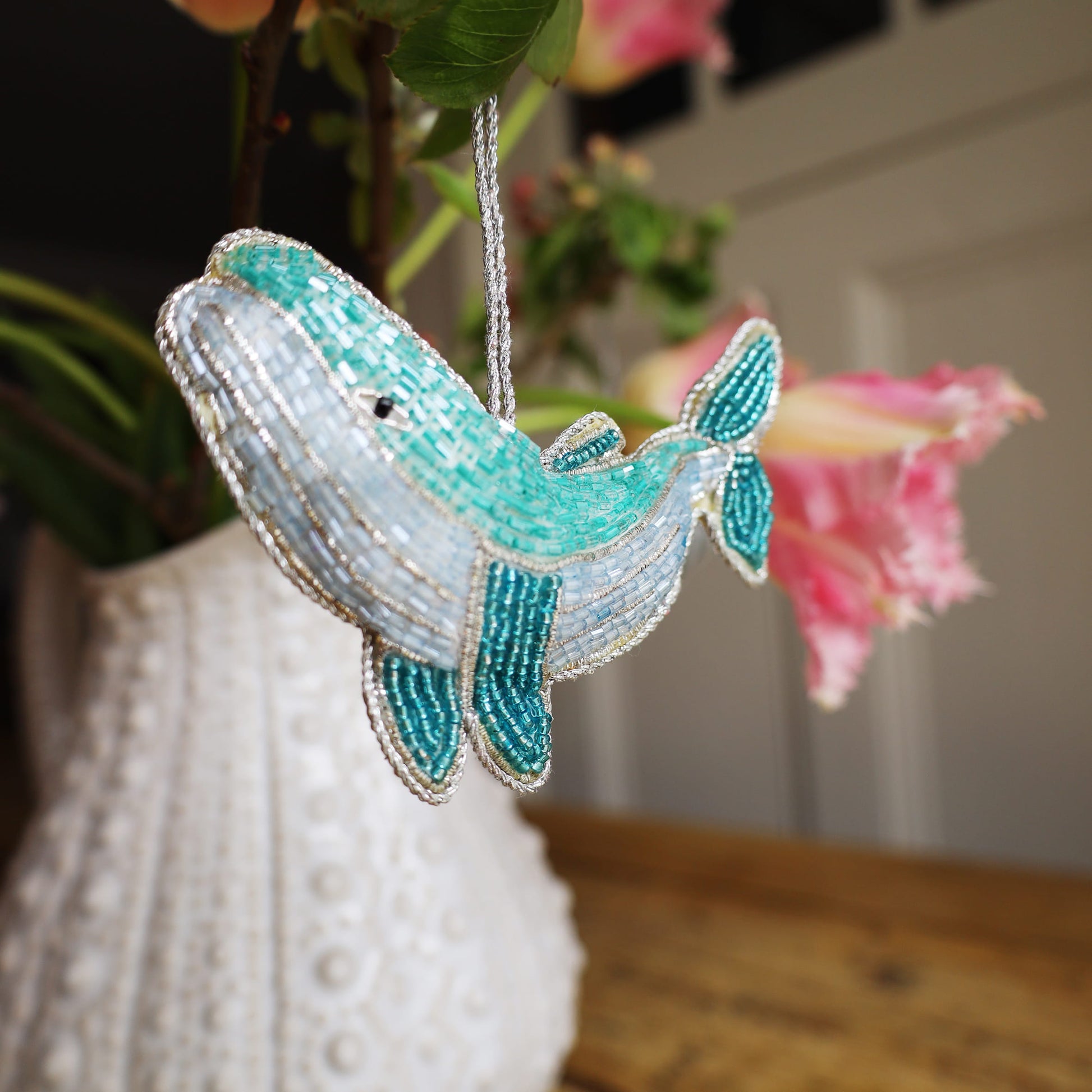 Decorative whale ornament with beaded details hanging near a white vase and pink flowers.