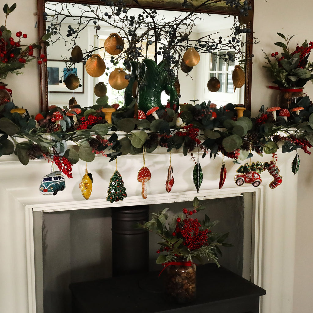 A festive fireplace mantel decorated with a garland of eucalyptus leaves, red berries, and small mushrooms, complemented by hanging beaded ornaments. The ornaments include a blue and white VW camper van, a yellow lemon, a Christmas tree, a mushroom, and a red car carrying a Christmas tree on its roof. Above the mantel, golden seashell ornaments hang from branches, reflecting in a mirror, while a green vase and additional holiday decor complete the cozy scene.