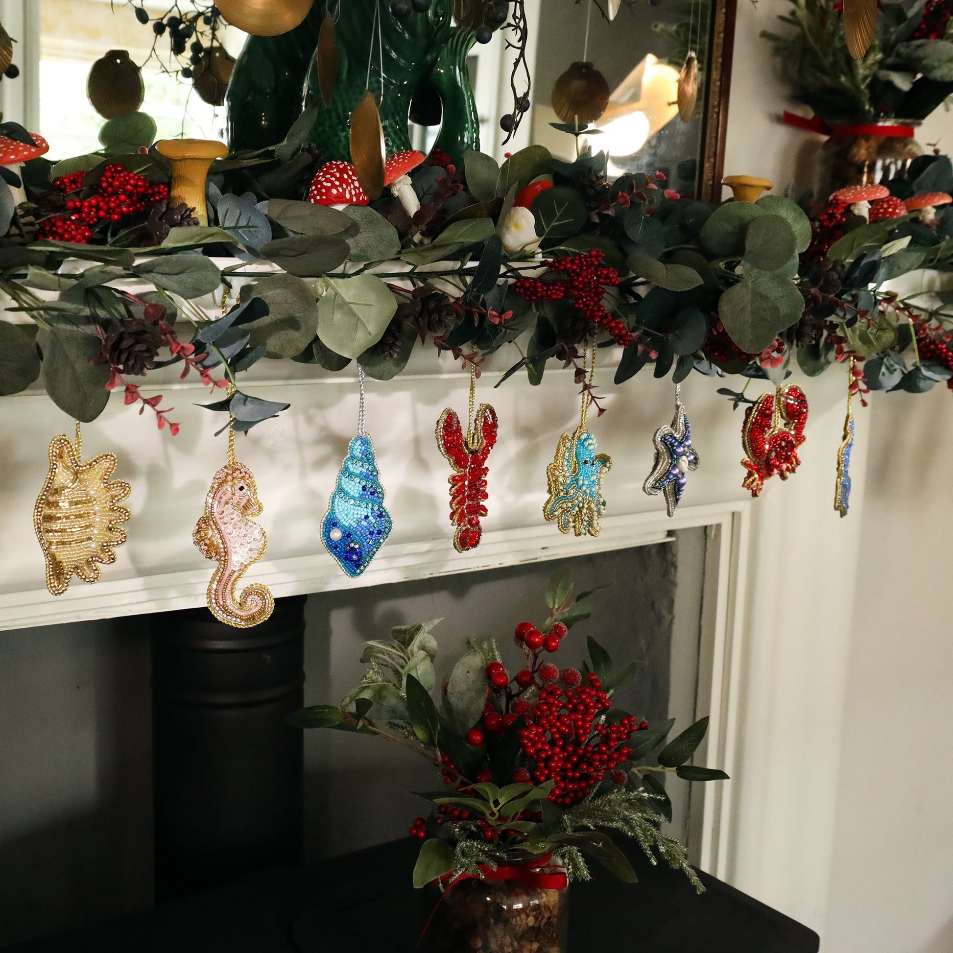 A festive garland draped over a mantel, adorned with beaded sea-themed ornaments. From left to right, there is a gold beaded shell, a pink seahorse, a turquoise shell, a red lobster, a blue octopus, a starfish with pearls, and another red crab. The garland itself is made of green leaves, red berries, and small mushrooms, adding a natural, woodland touch to the scene. Below the garland, a small bouquet of red berries sits on the mantel, enhancing the festive atmosphere.