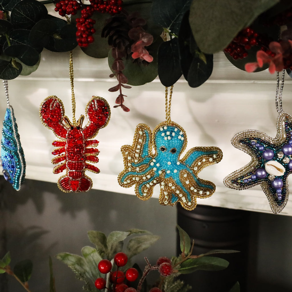 Festive beaded ornaments hanging from a mantel garland, featuring a bright red lobster, a turquoise octopus, and a purple starfish. The ornaments are intricately detailed with shimmering beads, surrounded by greenery and red berries, creating a cheerful holiday display with a coastal theme.