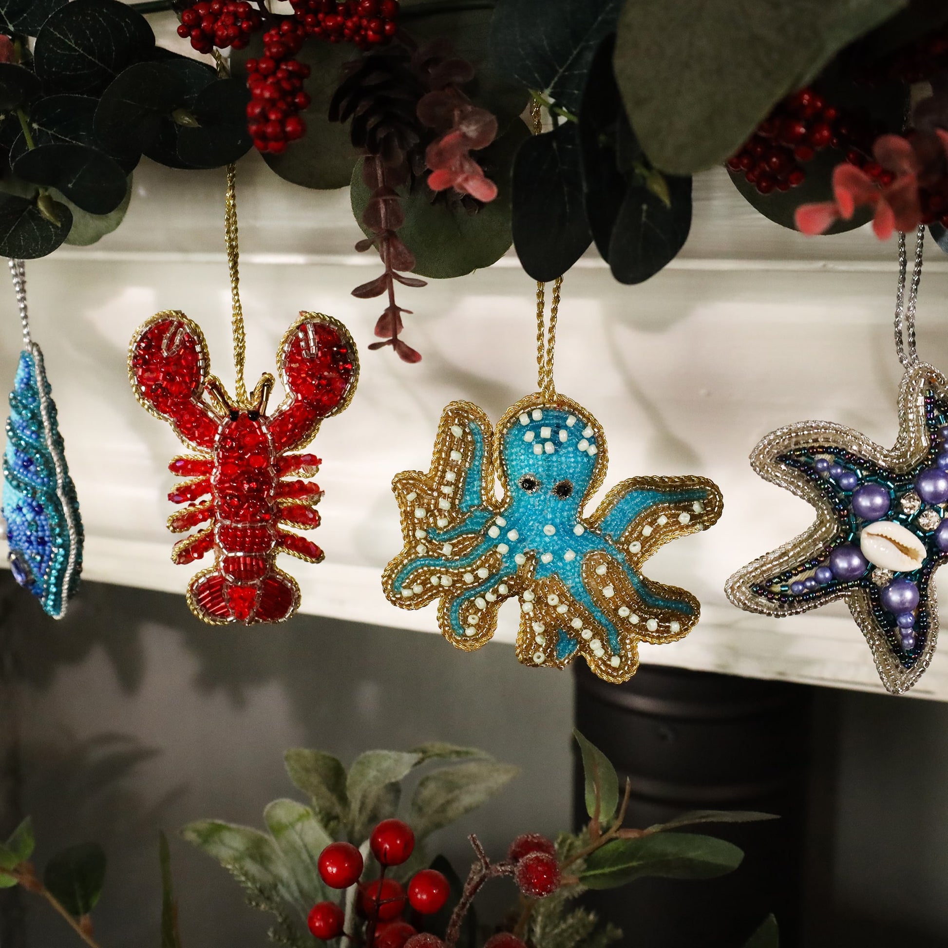 Festive beaded ornaments hanging from a mantel garland, featuring a bright red lobster, a turquoise octopus, and a purple starfish. The ornaments are intricately detailed with shimmering beads, surrounded by greenery and red berries, creating a cheerful holiday display with a coastal theme.
