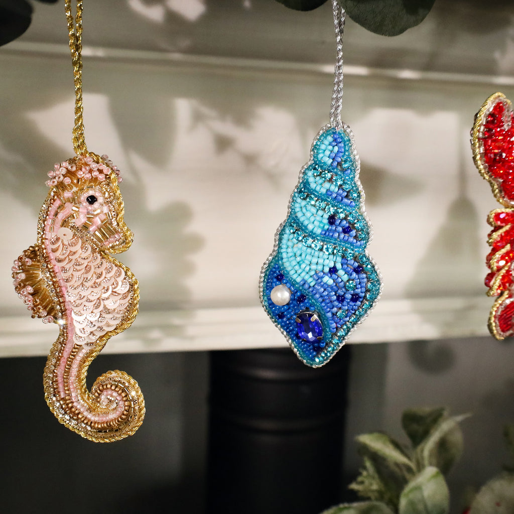 A close-up of two intricately beaded Christmas ornaments hanging from a garland. On the left is a pink and gold seahorse with shimmering sequins, and on the right is a turquoise beaded shell adorned with pearls and blue gemstones. Both ornaments are delicately crafted, adding a touch of oceanic elegance to the festive decor.