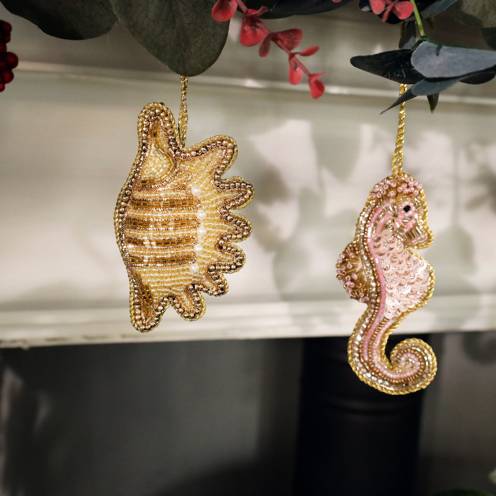 Close-up of two intricately beaded Christmas decorations hanging from a mantel garland. The left ornament resembles a gold shell adorned with pearl accents, while the right is a pink seahorse with sequins and beads. Both decorations have shimmering gold trim and hang from delicate gold strings.