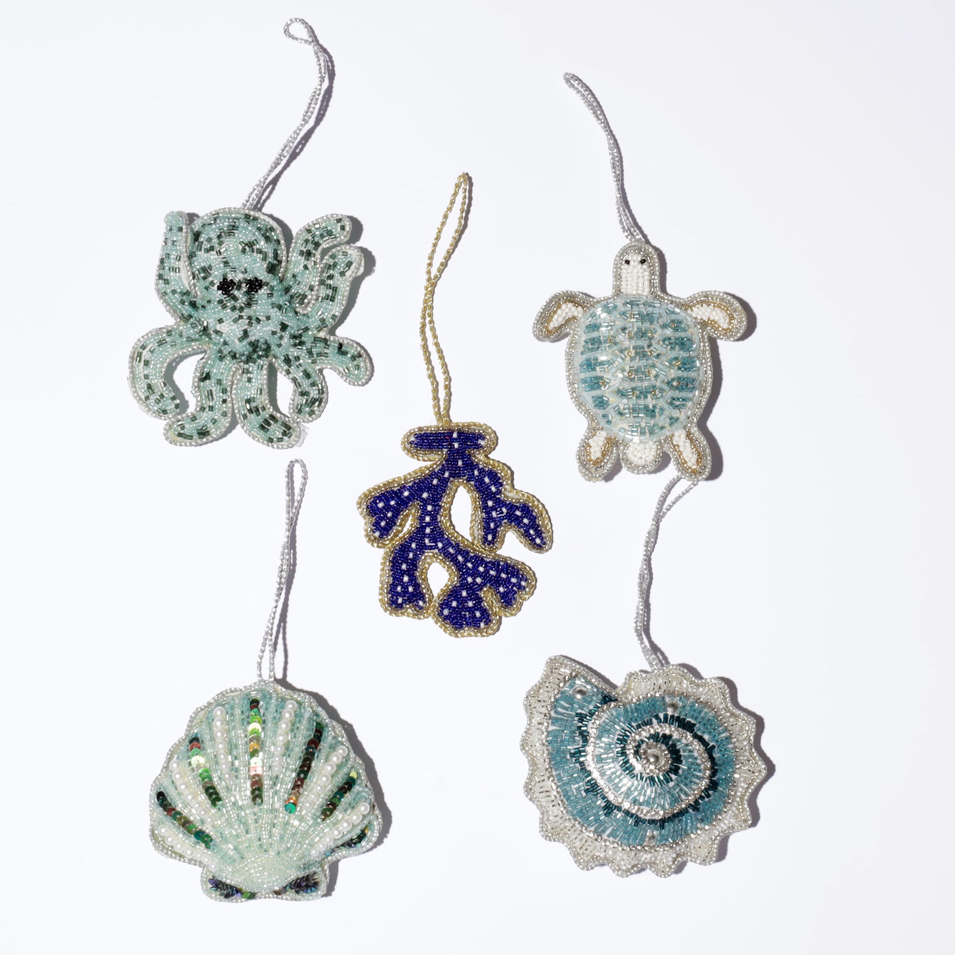 A collection of beautifully beaded ocean-themed decorations, arranged in a flat-lay on a white background. The ornaments include a light green beaded octopus, a sparkling blue and white turtle, a deep navy coral branch with gold trim, a shimmery sea shell, and a delicate spiral shell design. Each piece is intricately crafted with shimmering beads that capture the essence of the ocean and its vibrant marine life.