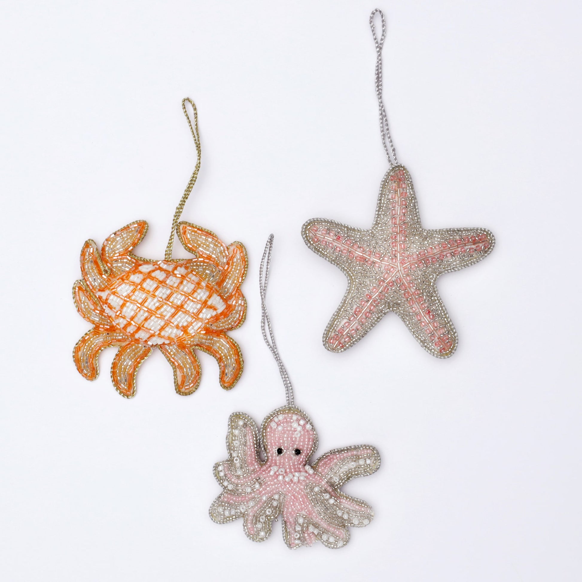 A delightful trio of beaded marine-themed ornaments featuring a vibrant orange crab, a soft pink starfish, and an adorable pink octopus. Each ornament is intricately crafted with shimmering beads, detailed accents, and metallic threading, creating a playful and elegant under-the-sea aesthetic. The ornaments rest on a white background, showcasing their vibrant colours and exquisite craftsmanship, perfect for adding a touch of seaside charm to any décor.