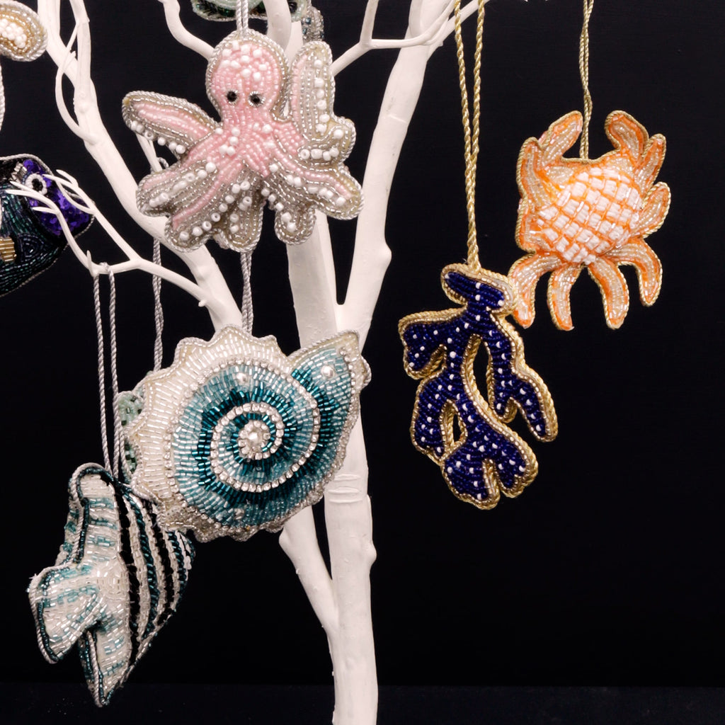 A collection of intricately beaded ocean-themed ornaments hanging on a white display tree against a black background. Featured designs include a pink octopus, a teal and white spiral shell, a navy coral branch with gold trim, and an orange crab. Each piece is detailed with shimmering beads, creating a vibrant and elegant underwater-inspired display.