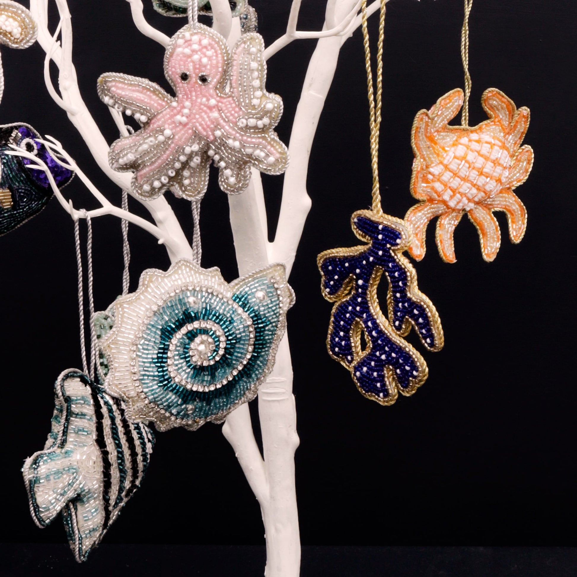 A collection of intricately beaded ocean-themed ornaments hanging on a white display tree against a black background. Featured designs include a pink octopus, a teal and white spiral shell, a navy coral branch with gold trim, and an orange crab. Each piece is detailed with shimmering beads, creating a vibrant and elegant underwater-inspired display.