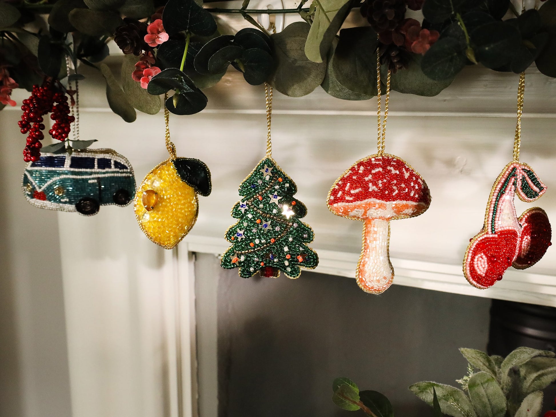 A festive garland featuring five colorful beaded ornaments hanging from a lush eucalyptus arrangement. The decorations include a teal van, a bright yellow lemon, a green Christmas tree adorned with stars, a red and white mushroom, and a pair of red cherries. Each ornament is intricately designed with beads and sequins, creating a vibrant and whimsical holiday display.