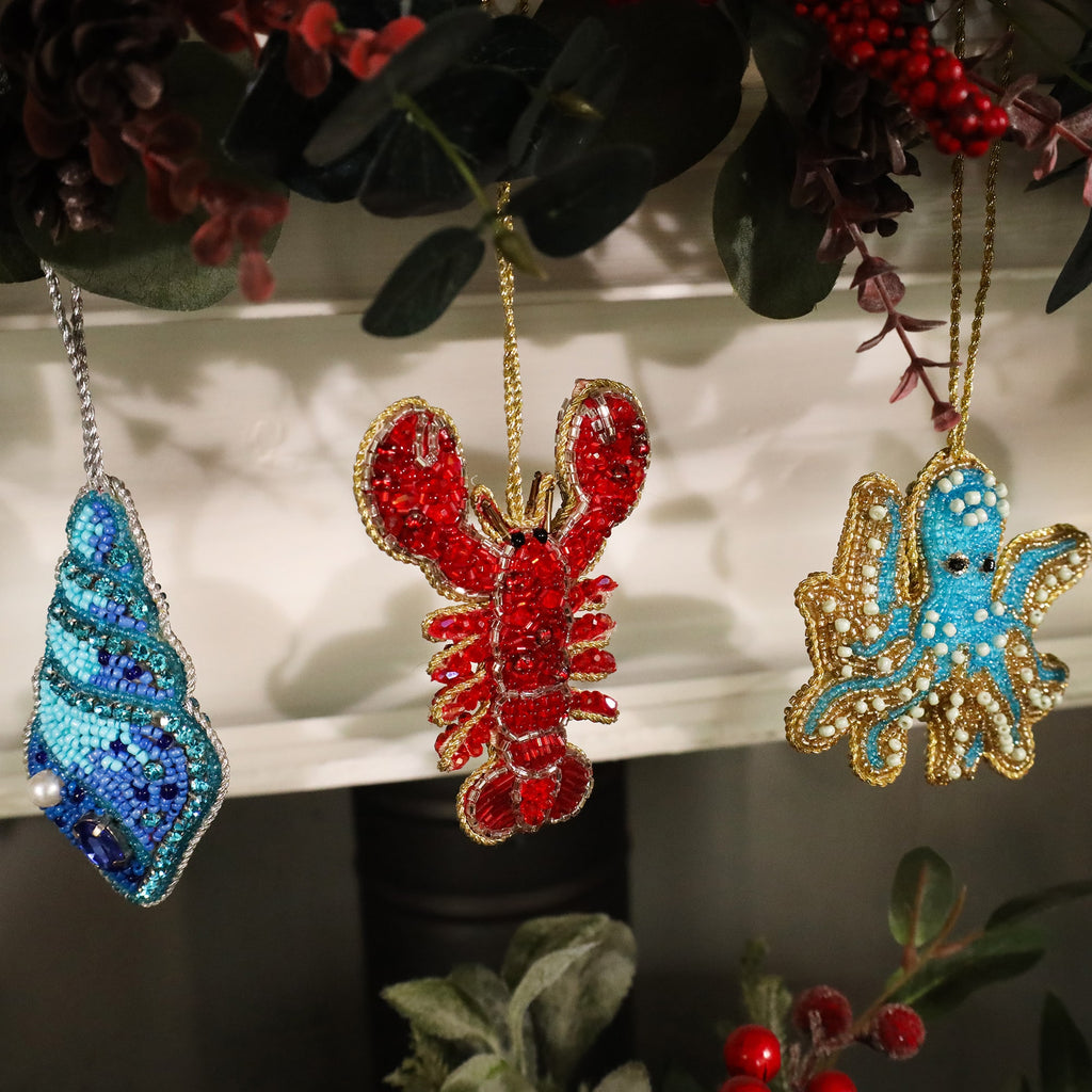 A trio of colorful beaded ornaments hanging on a festive garland. From left to right: a blue seashell with intricate beadwork and a pearl detail, a bright red lobster with gold accents, and a turquoise octopus outlined in gold. The ornaments are beautifully detailed and give a coastal theme to the holiday decor.