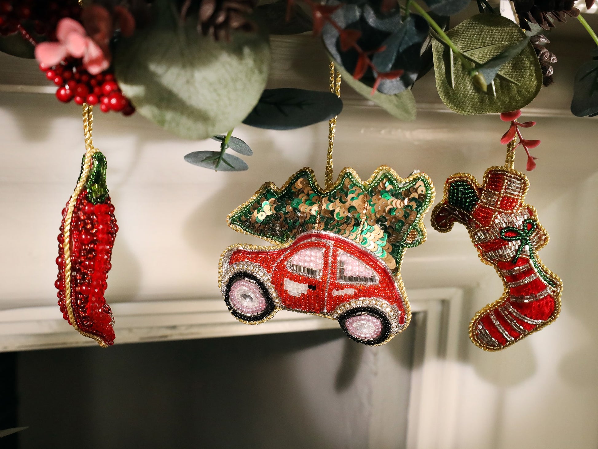 Three festive beaded ornaments hang in a row. On the left is a red chili pepper, in the center a red car carrying a sequined Christmas tree on its roof, and on the right is a red and white striped Christmas stocking with silver beading and a green bow. The ornaments are outlined with gold beads, suspended from greenery with a cozy holiday feel.
