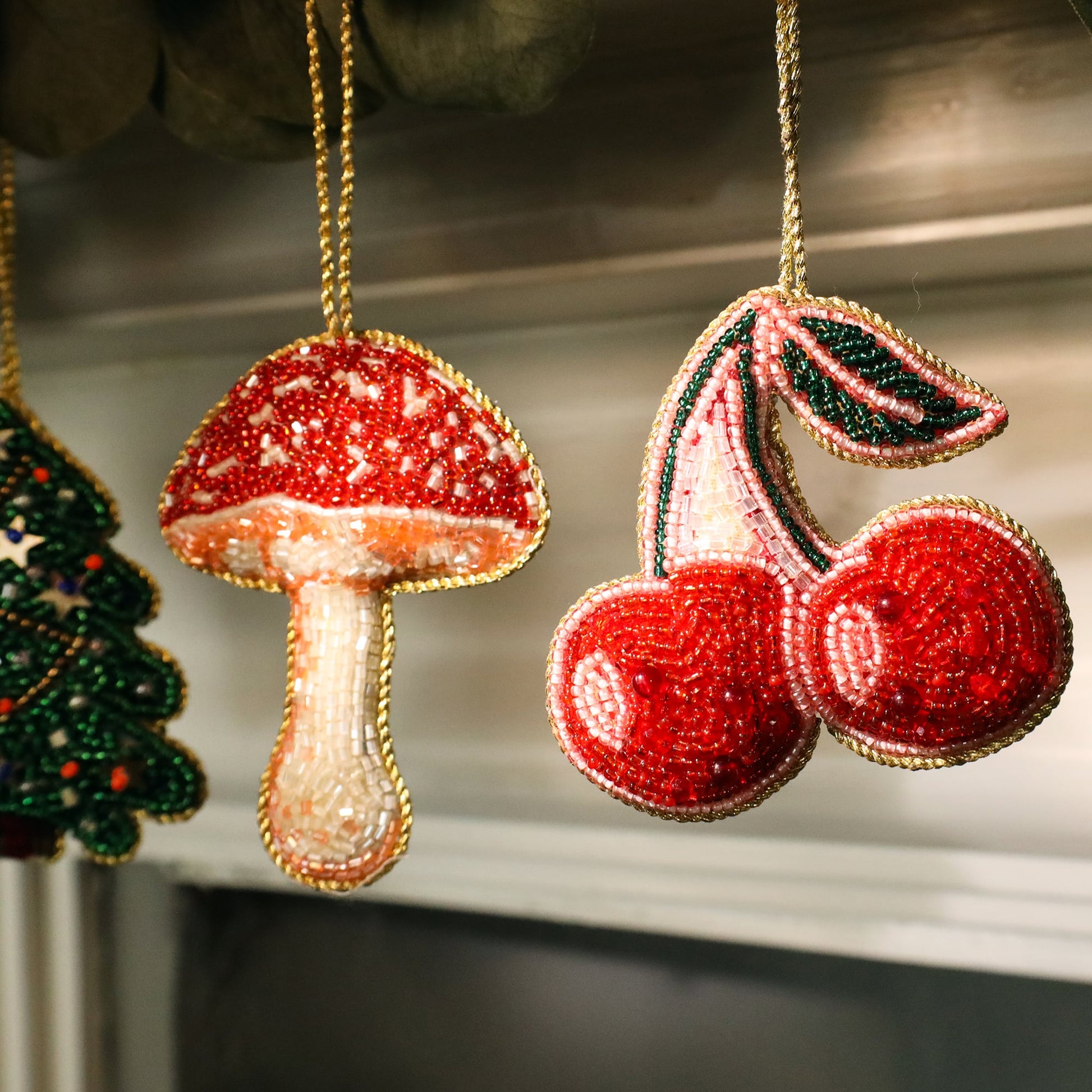 Close-up of two beaded Christmas ornaments hanging from a garland. One ornament is a red and white mushroom with intricate beading, and the other is a pair of bright red cherries with green leaves. Both are outlined in gold beads, adding a festive and sparkling touch to the decorations.