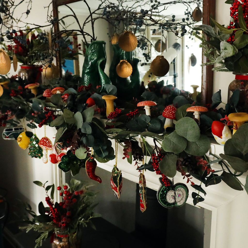 A festive mantle display adorned with a garland of eucalyptus leaves, red berries, and whimsical mushroom decorations. Hanging from the garland are various beaded ornaments, including a green apple, chili pepper, and leaf, all intricately detailed. Golden shell decorations hang from branches above, while green ceramic vases and other holiday accents complete the cozy, nature-inspired scene.