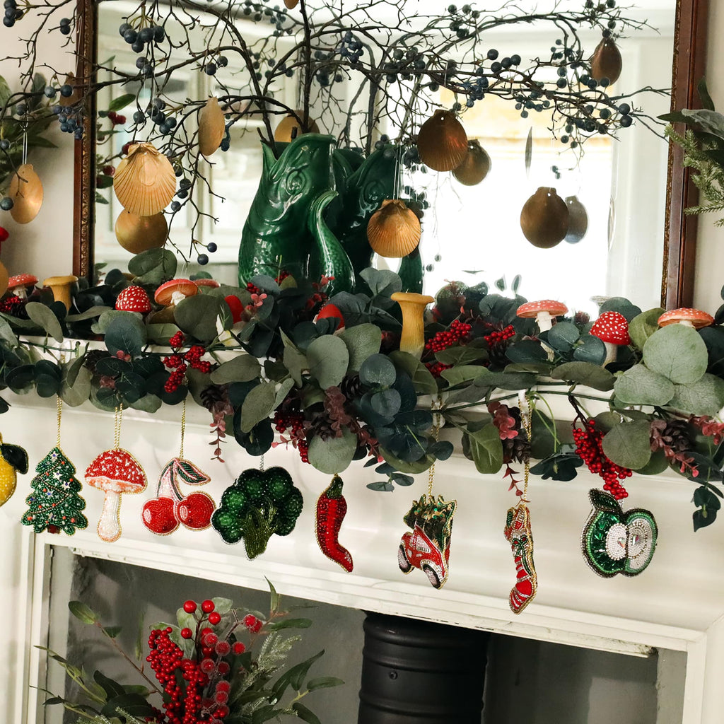 A festive fireplace mantel decorated with lush greenery, red berries, and beaded ornaments. The beaded decorations include a Christmas tree, mushroom, cherries, broccoli, chili pepper, car with a Christmas tree, a stocking, and an apple slice. Above the mantel, branches with golden seashell ornaments and eucalyptus leaves add a natural touch, creating a warm and whimsical holiday display.