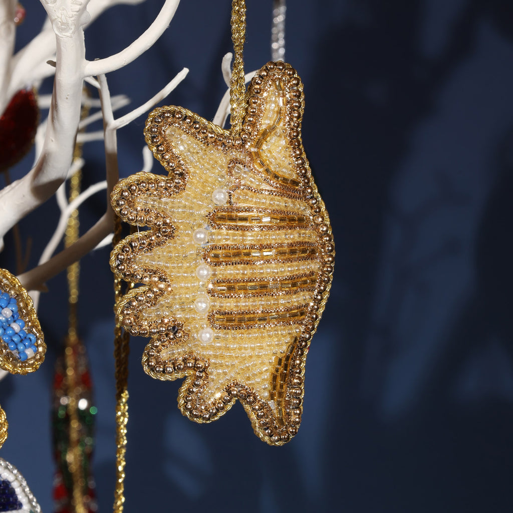A close-up of a beaded golden seashell ornament, intricately decorated with shimmering gold beads and pearl accents. The ornament hangs from a white branch against a dark background, reflecting light with its metallic and detailed beadwork.