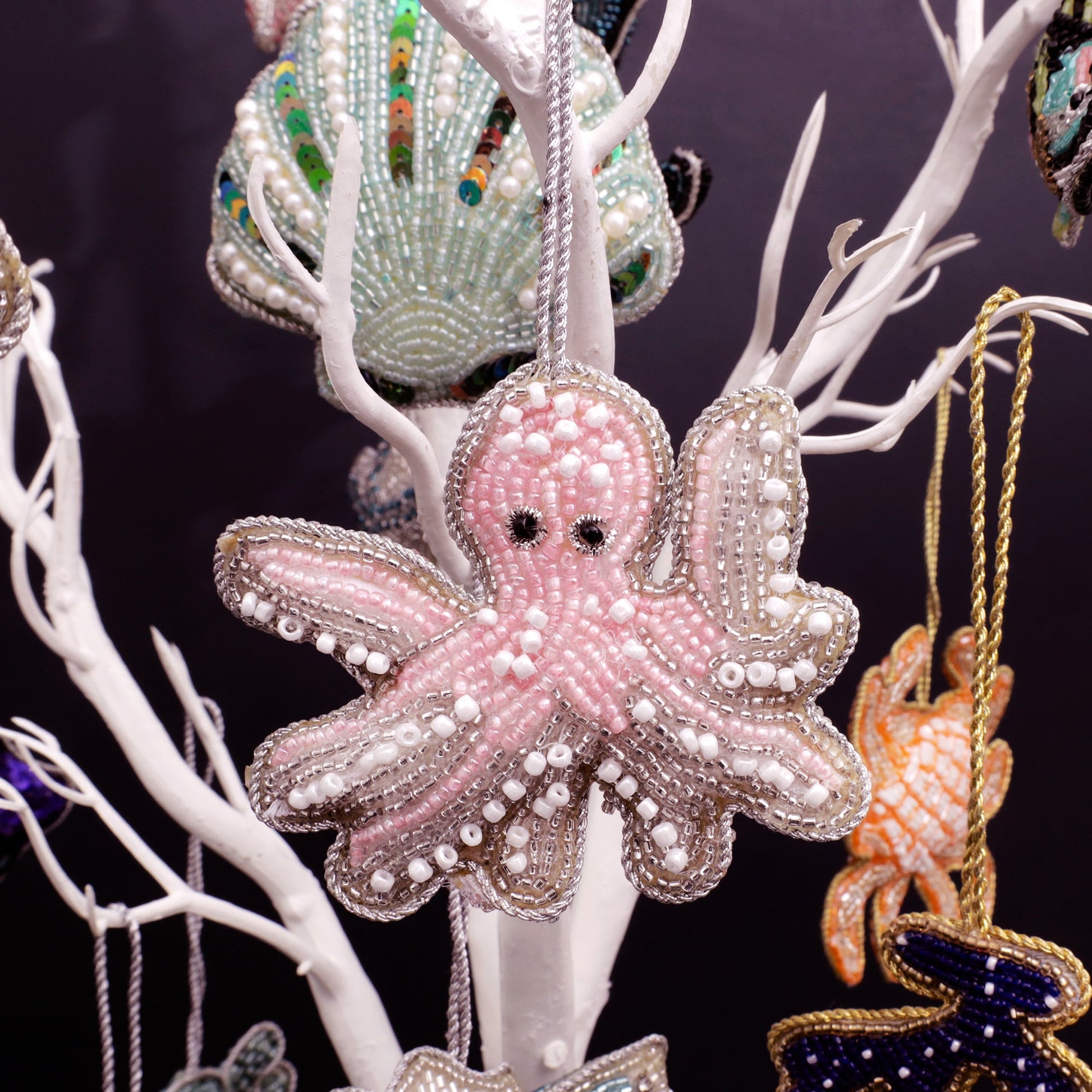 A whimsical pink octopus ornament, meticulously crafted with shimmering beads and pearl accents, hanging gracefully on a white decorative tree. The octopus features intricate pink and white beadwork, highlighted by silver-edged detailing and expressive black beaded eyes. Surrounding the octopus are other beaded marine-themed decorations, adding a playful underwater charm to the scene.