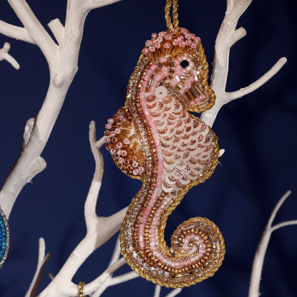 A close-up of a beautifully beaded pink seahorse ornament hanging on a white branch display. The seahorse is intricately crafted with shimmering beads, sequins, and gold accents, giving it a delicate and elegant appearance. The details highlight the scales, curling tail, and intricate body of the seahorse, creating a stunning ocean-inspired decoration.