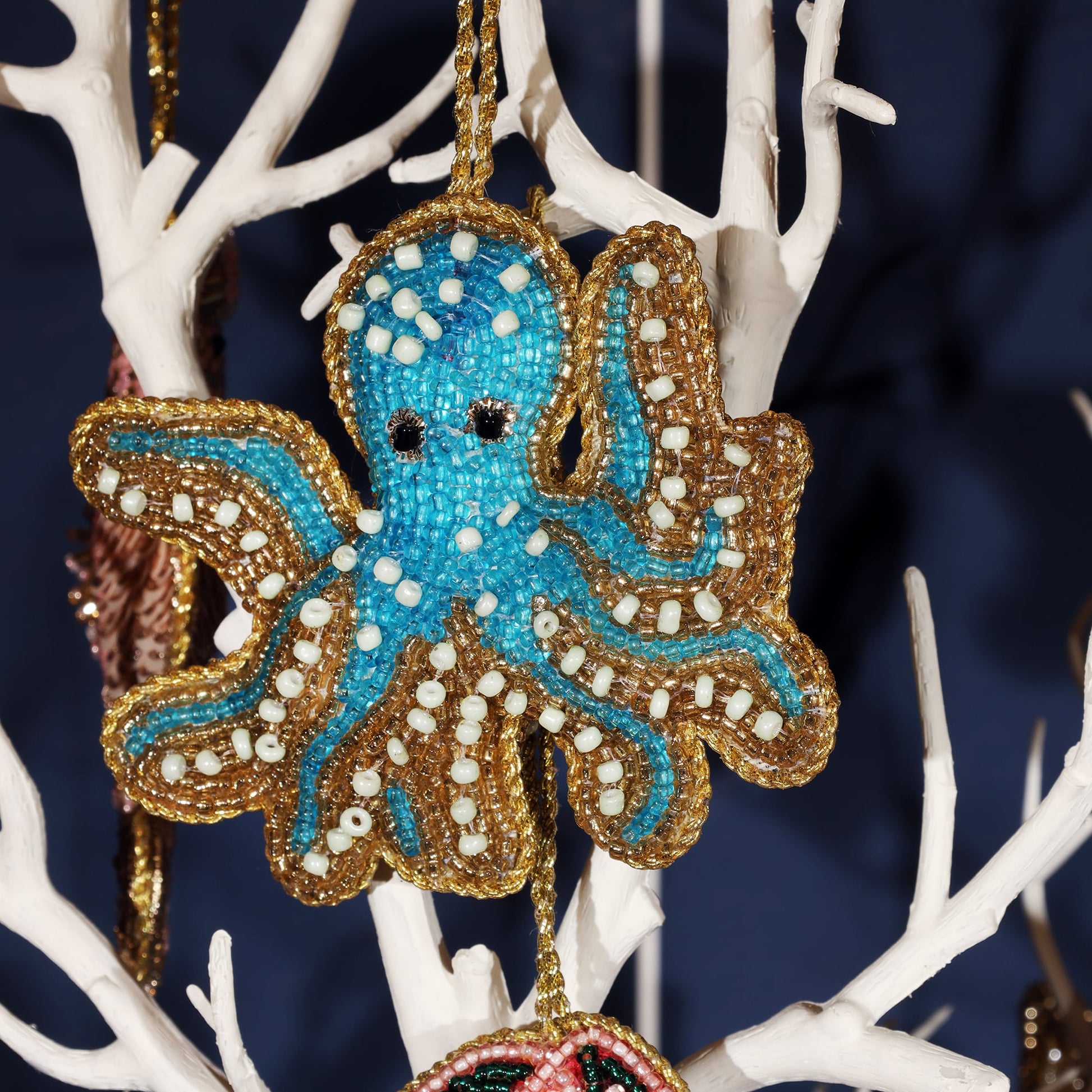 Close-up of a turquoise beaded octopus ornament hanging on a white tree branch. The octopus is intricately detailed with blue, gold, and white beads, with black bead eyes, and outlined in gold beadwork, giving it a festive and playful ocean-themed appearance.