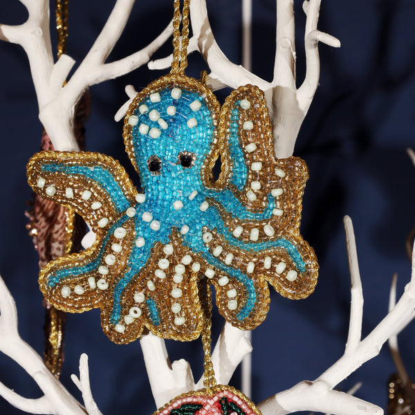 Close-up of a turquoise beaded octopus ornament hanging on a white tree branch. The octopus is intricately detailed with blue, gold, and white beads, with black bead eyes, and outlined in gold beadwork, giving it a festive and playful ocean-themed appearance.