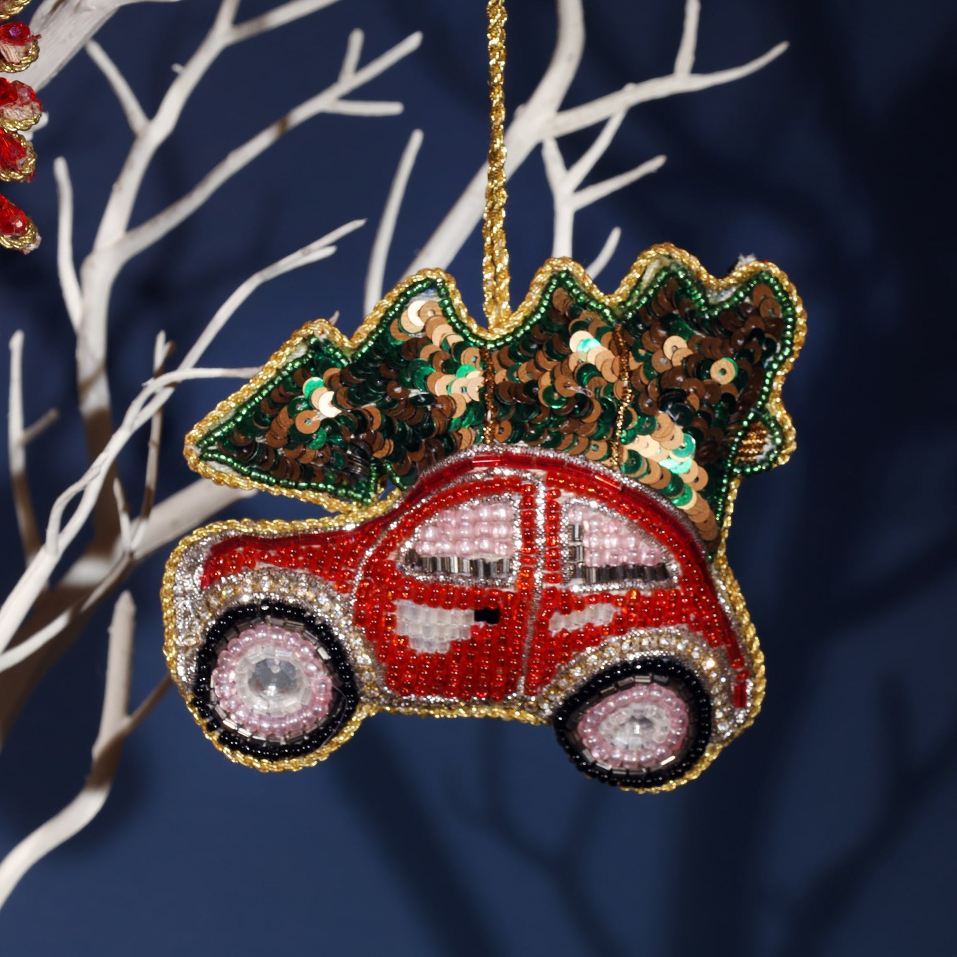 A detailed, beaded ornament of a red car carrying a Christmas tree on its roof. The car is intricately designed with red, silver, and black beads, and the windows are adorned with shimmering pink beads. The tree on top is decorated with green and brown sequins, giving it a festive sparkle. The ornament is bordered with gold beads, adding an elegant touch to the playful holiday theme.