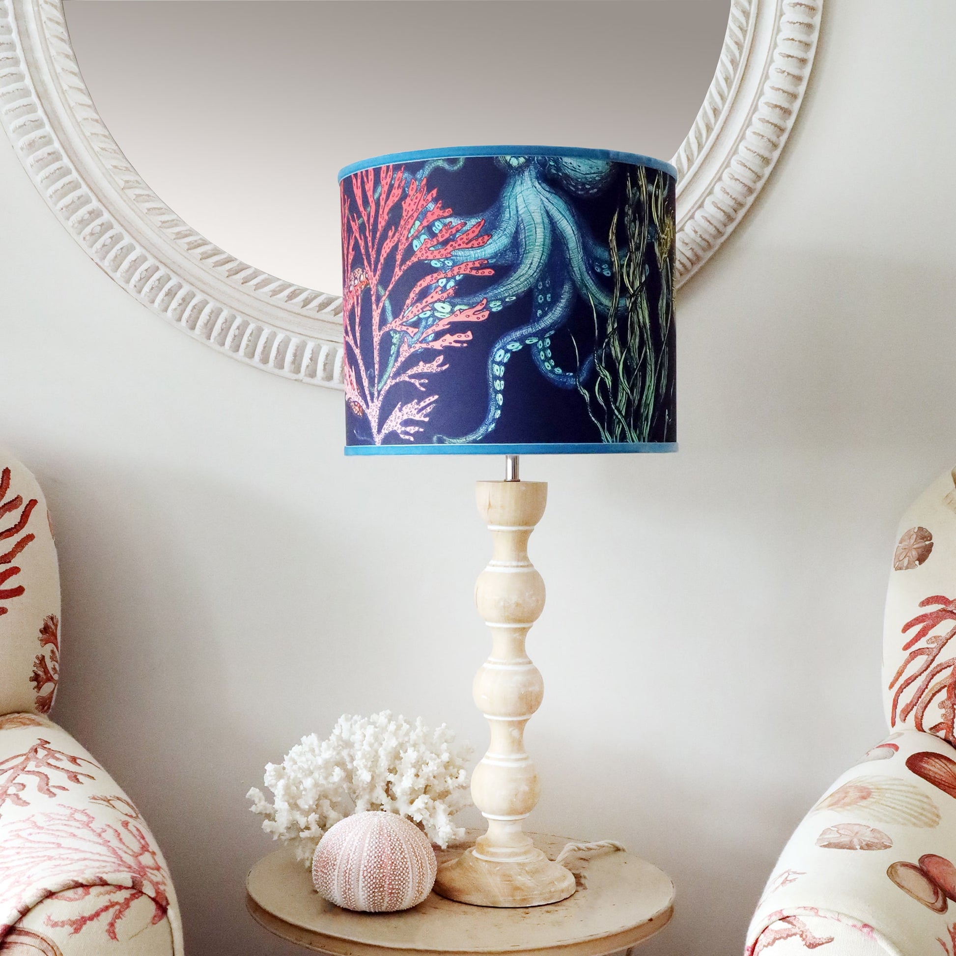 The image shows a coastal-themed interior with a white, circular mirror featuring a rope-like textured frame on the wall. In front of the mirror, there is a wooden table lamp with a curvy, light wood base and a lampshade decorated with a vibrant marine design, including an octopus and coral in shades of blue, pink, and green. The lamp sits on a small round table, which also holds a piece of white coral and a pink sea urchin. On either side of the table, there are upholstered armchairs with a coral and seash