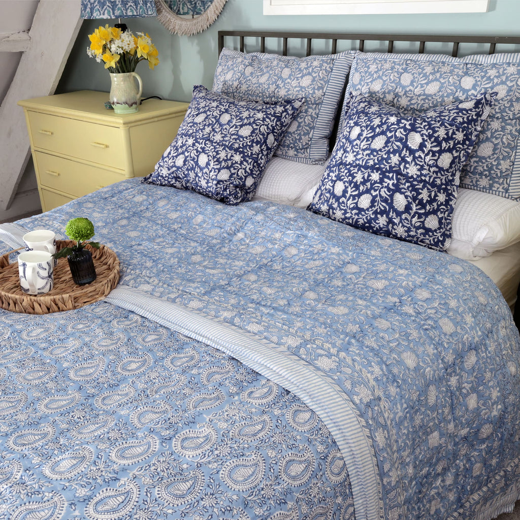 A charming bedroom setup featuring a bed dressed with light blue and white block-printed bedding with intricate floral and paisley designs. Dark blue cushions with floral and shell patterns are arranged against striped and patterned pillows. A woven tray on the bed holds two mugs and a small vase with a green flower. A yellow bedside table with a pitcher of daffodils adds a cheerful touch, complementing the cozy, rustic decor.