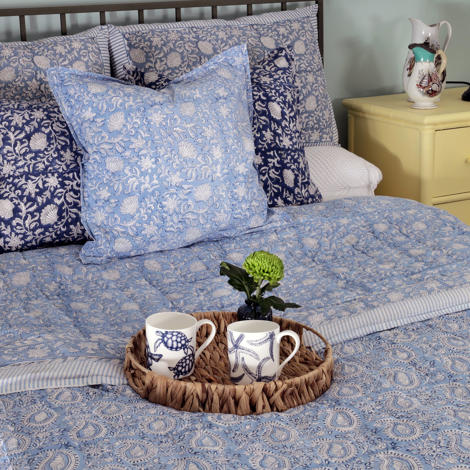 Bedroom setting featuring a bed with light blue and navy floral block print cushions and a matching quilt. A wicker tray on the bed holds two nautical-themed mugs and a small vase with a green flower, adding a cozy and decorative touch. A yellow nightstand with a pitcher completes the inviting scene.